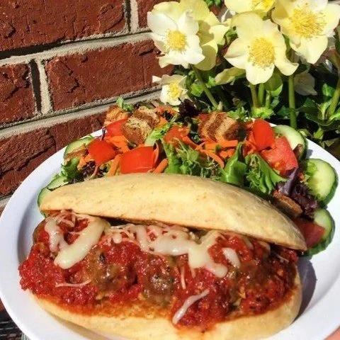 A plate with a meatball sub sandwich topped with melted cheese, accompanied by a side salad with cucumbers, tomatoes, and greens, and a bouquet of white and yellow flowers in the background.