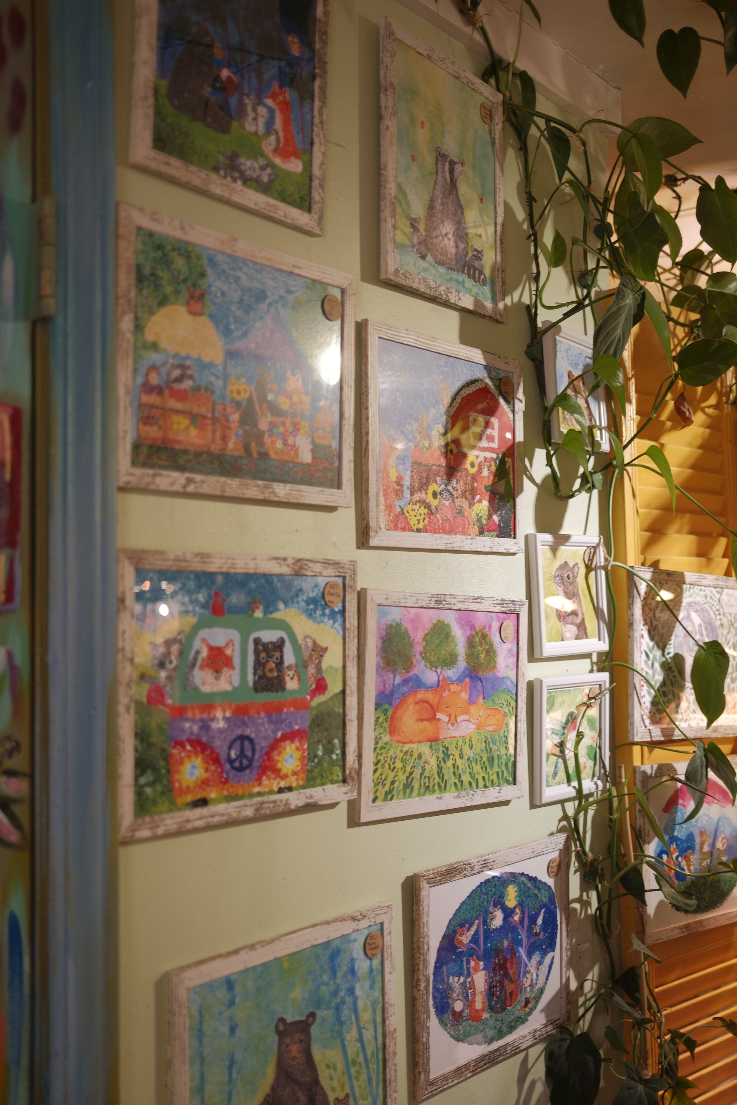 A collection of framed animal-themed artwork displayed on a green wall, with green leafy plants intertwined among the frames.