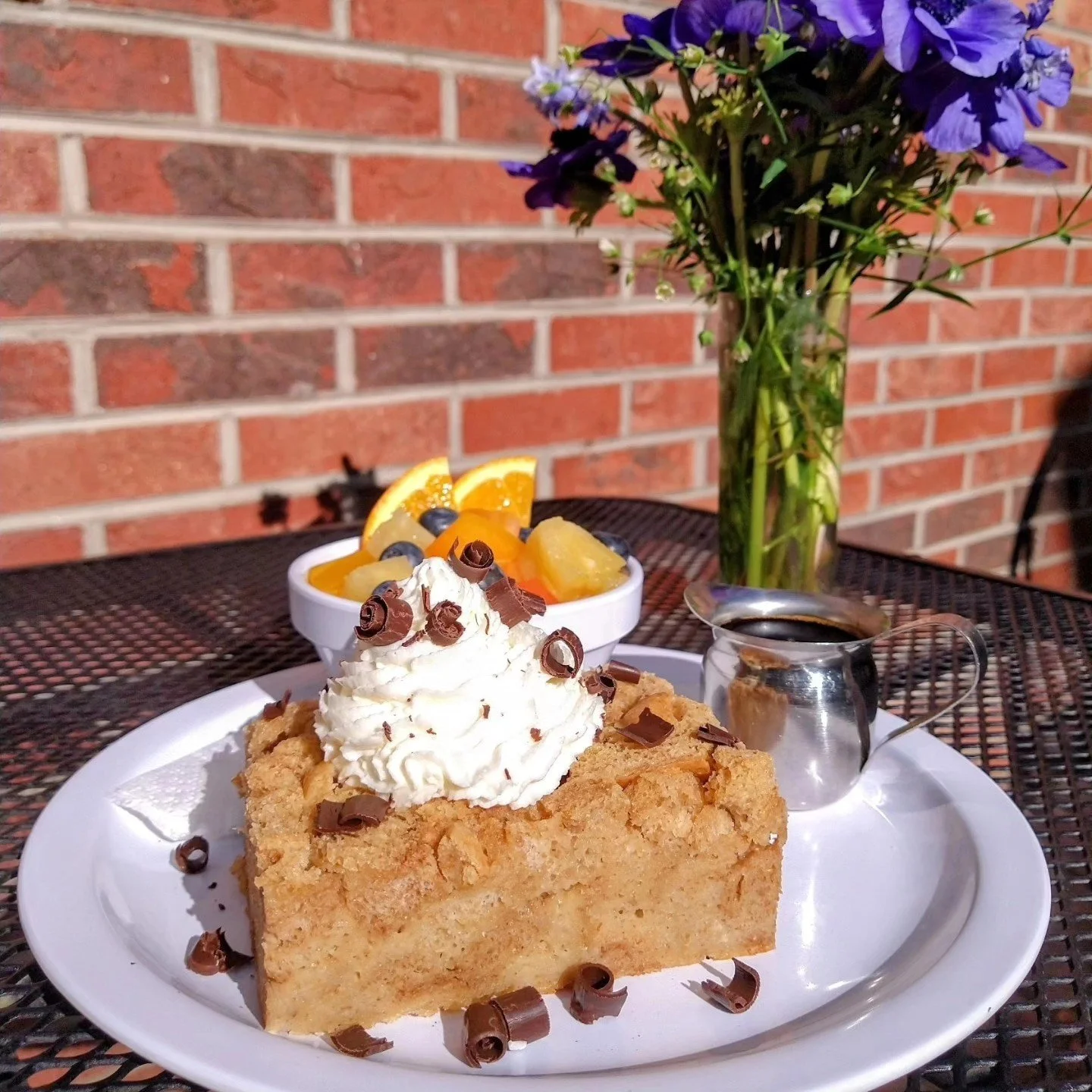 A slice of pie topped with whipped cream and chocolate curls, served with fruit and a side of syrup on a white plate, with a bouquet of purple flowers and a container of syrup on a dark table against a brick wall