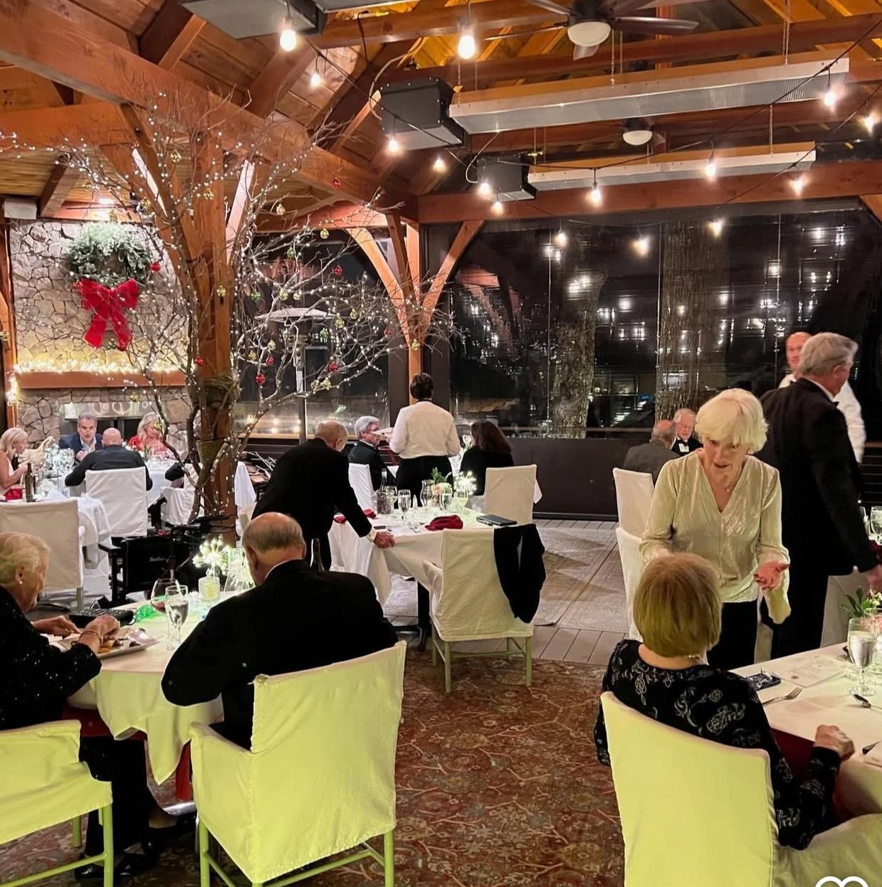 A festive indoor dining scene with people seated at white-draped tables, decorated with candles and glasses, in a warmly lit restaurant with wooden beams. Christmas decorations include a large wreath with a red bow and twinkling lights on leafless tr