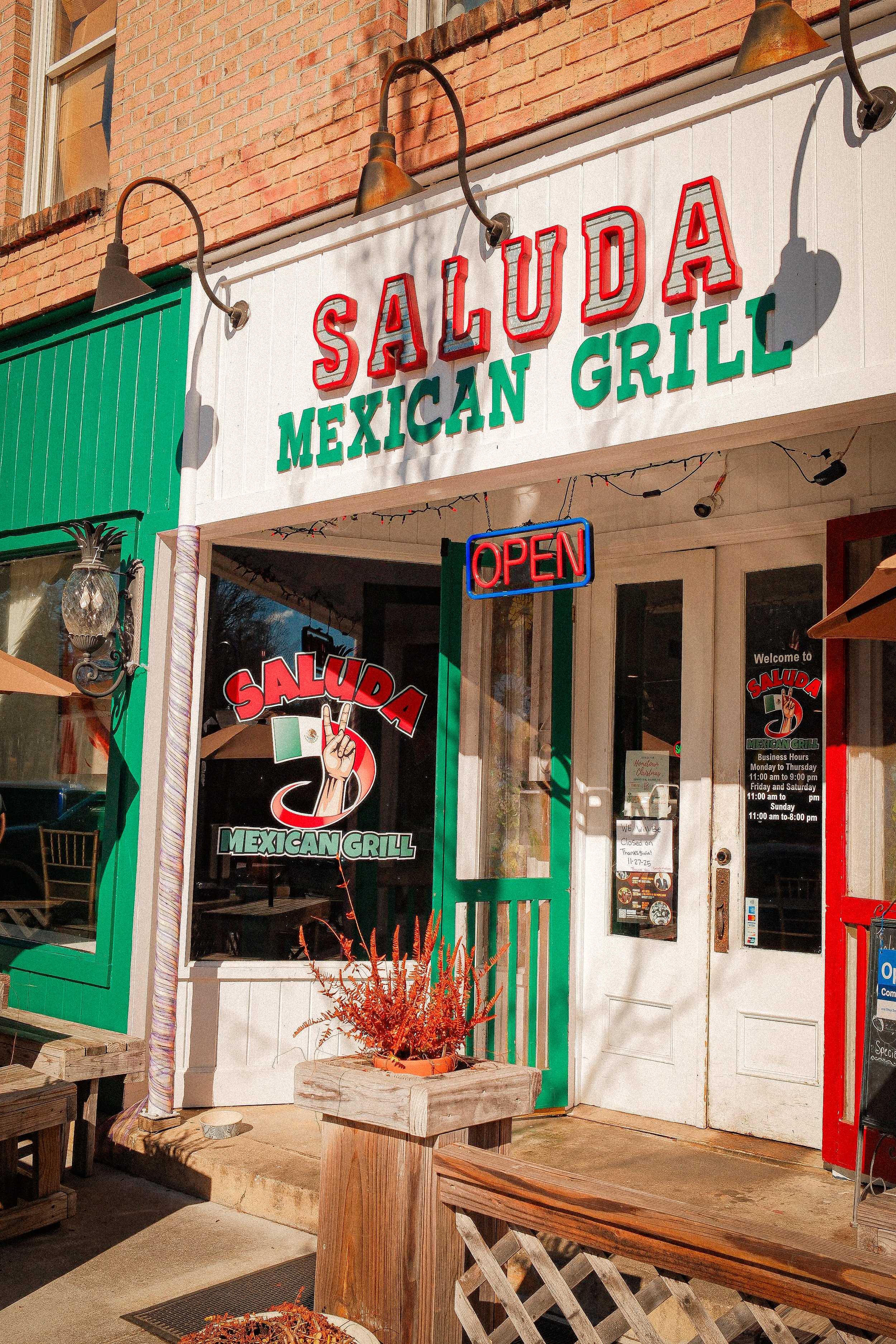 The photo shows the exterior of a Mexican restaurant called Saluda Mexican Grill with a sign saying 'Open' hanging below the restaurant's name. The entrance features a green door with a window, a white door, and some decorative outdoor lighting. There is a wooden bench and a planter with orange plants outside.