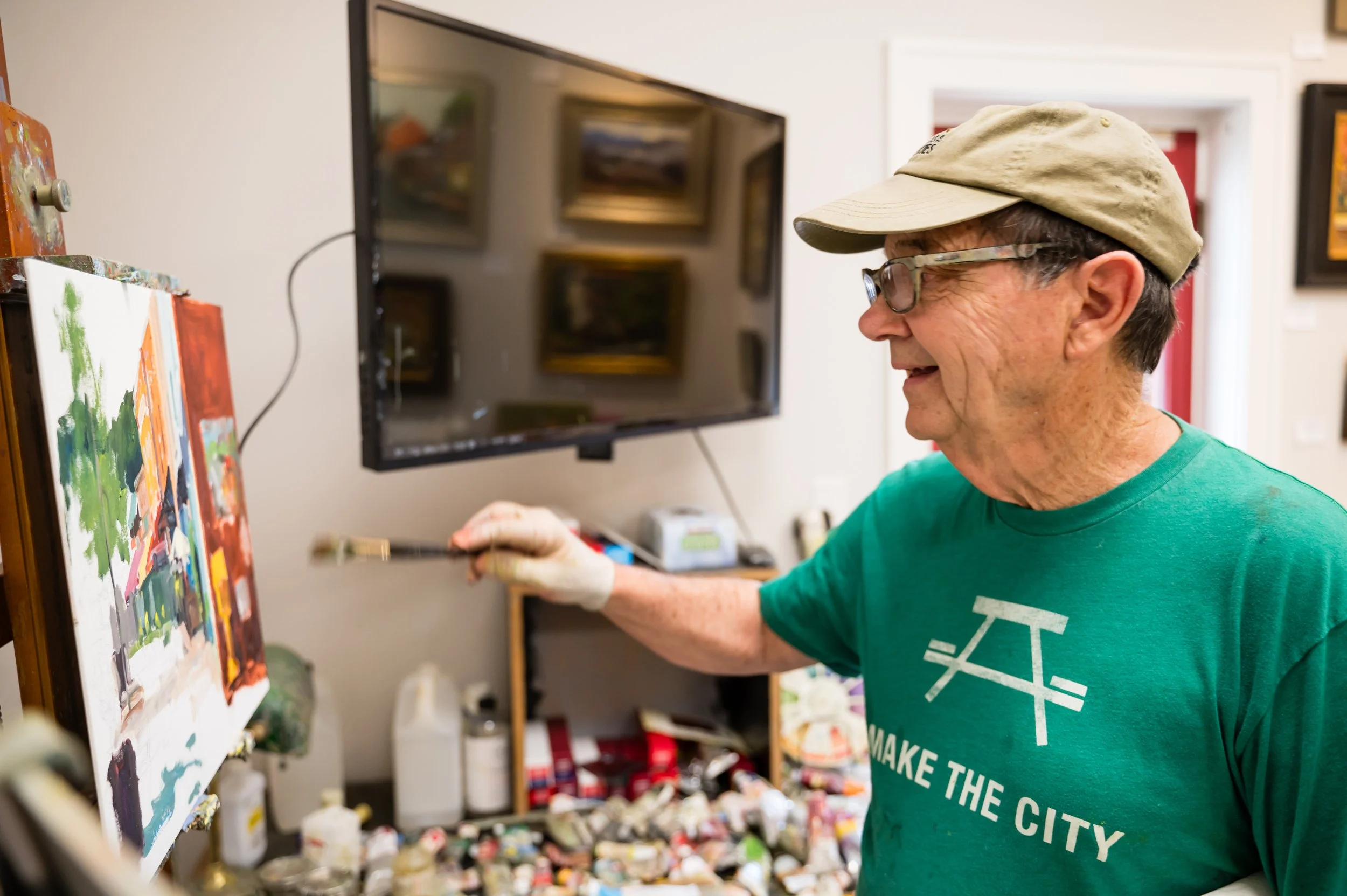 An older man wearing glasses, a beige cap, and a green T-shirt is smiling while painting on a canvas in an art studio. The studio has paintings on the wall and art supplies below a shelf.