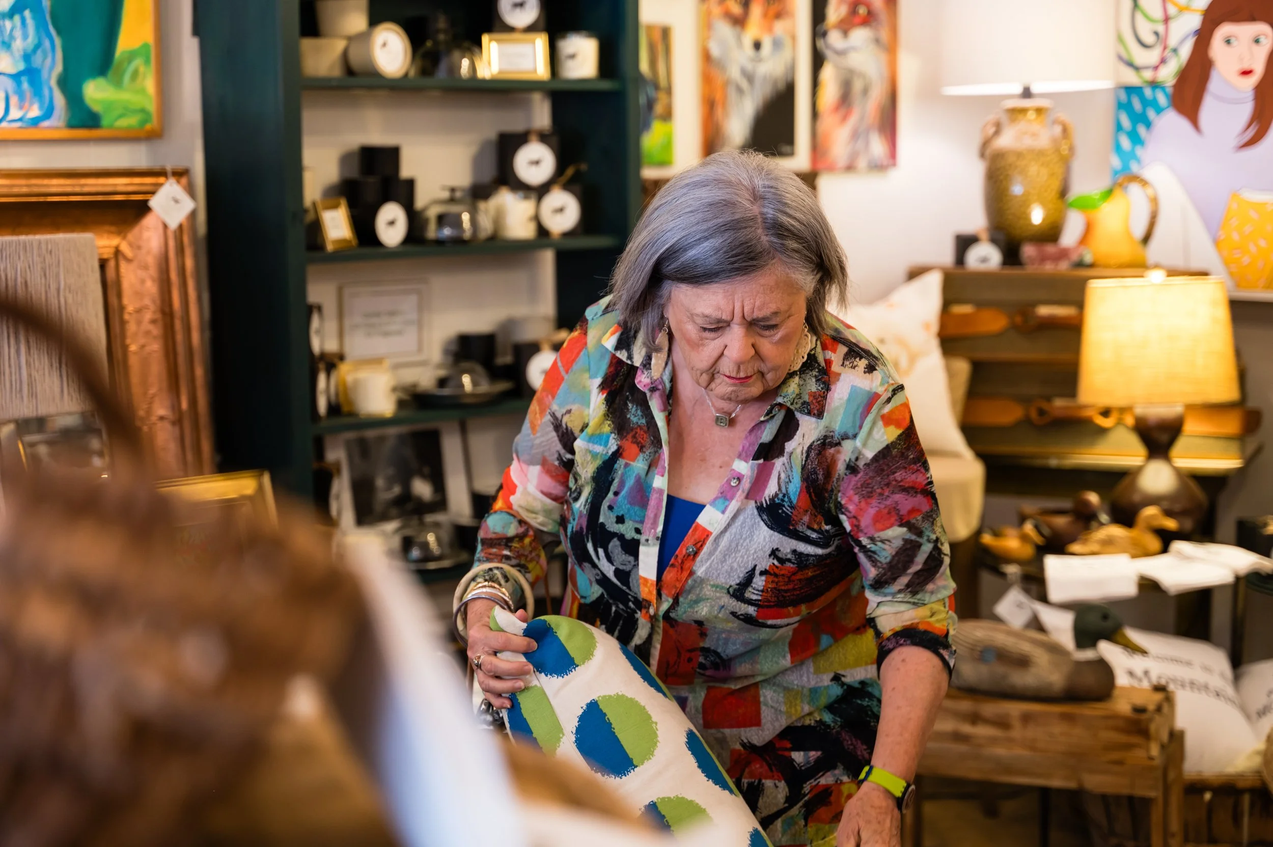 An elderly woman with gray hair wearing a multicolored shirt is examining a colorful piece of art or fabric at an art or craft store. The background shows shelves with clocks, picture frames, and various decorative items, with vibrant paintings on th