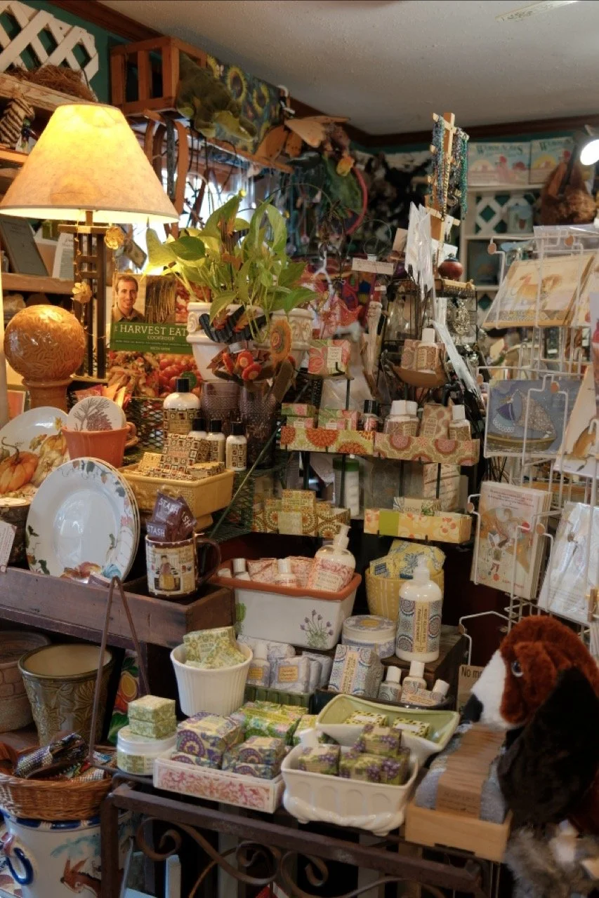 Interior of a gift shop filled with decorative items, plants, candles, ceramics, greeting cards, and a stuffed dog toy in the foreground.