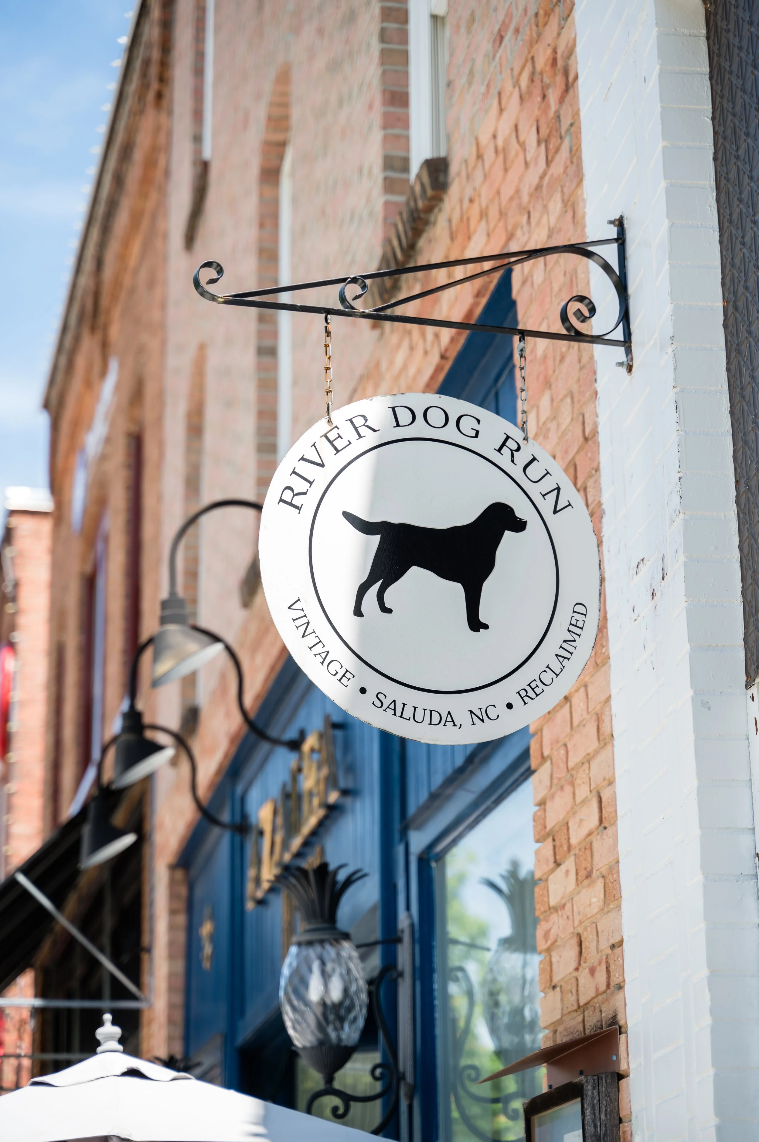 A black and white circular sign hanging from a black wrought iron bracket attached to a brick wall. The sign reads "River Dog Run" with a silhouette of a dog in the center and additional text around the circle. The background features a window with a blue frame and a street scene with lights.