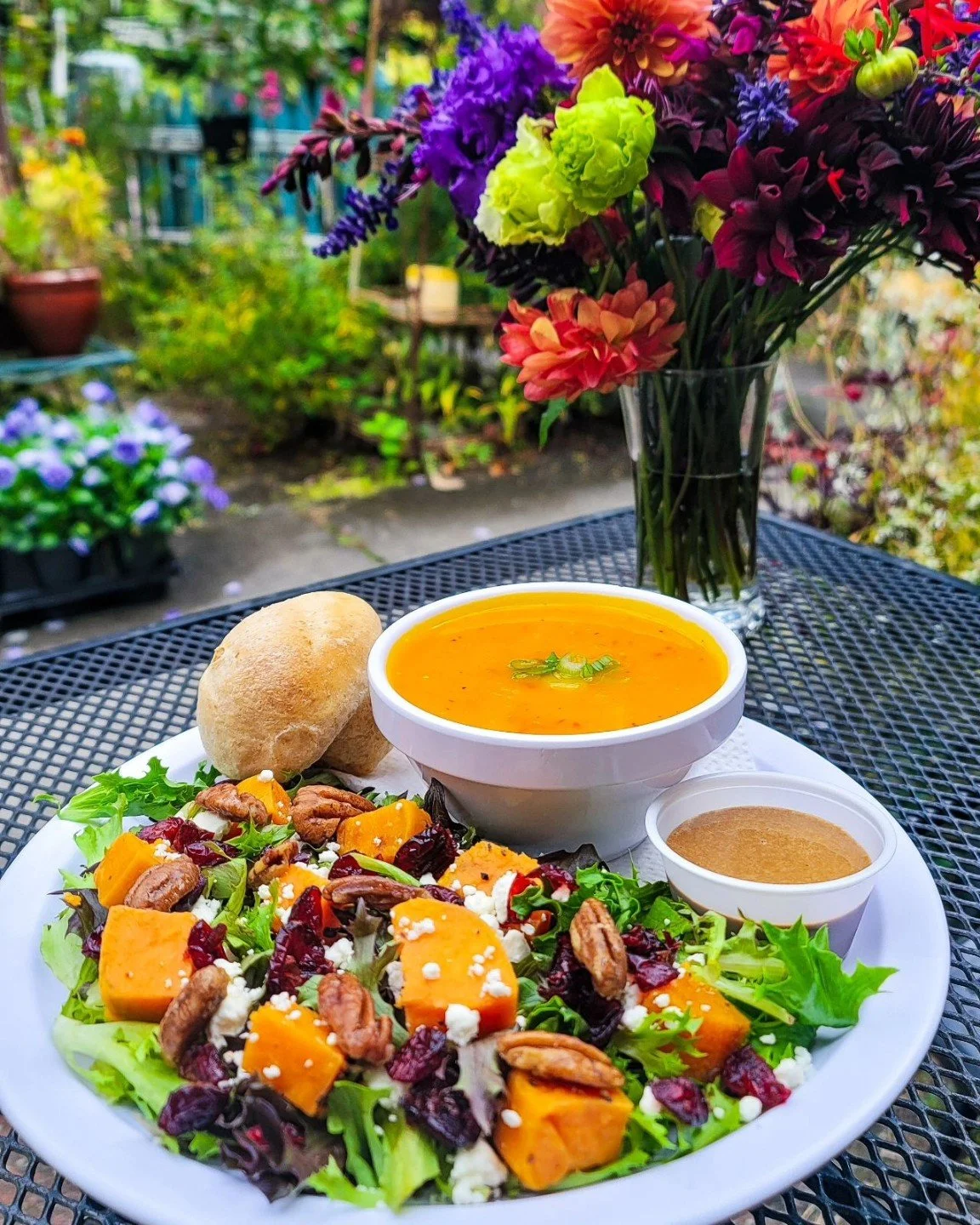 A colorful outdoor meal featuring a salad with roasted butternut squash, nuts, dried cranberries, and feta cheese, served with soup and bread, on a black metal table in a garden with vibrant flowers in a glass vase.