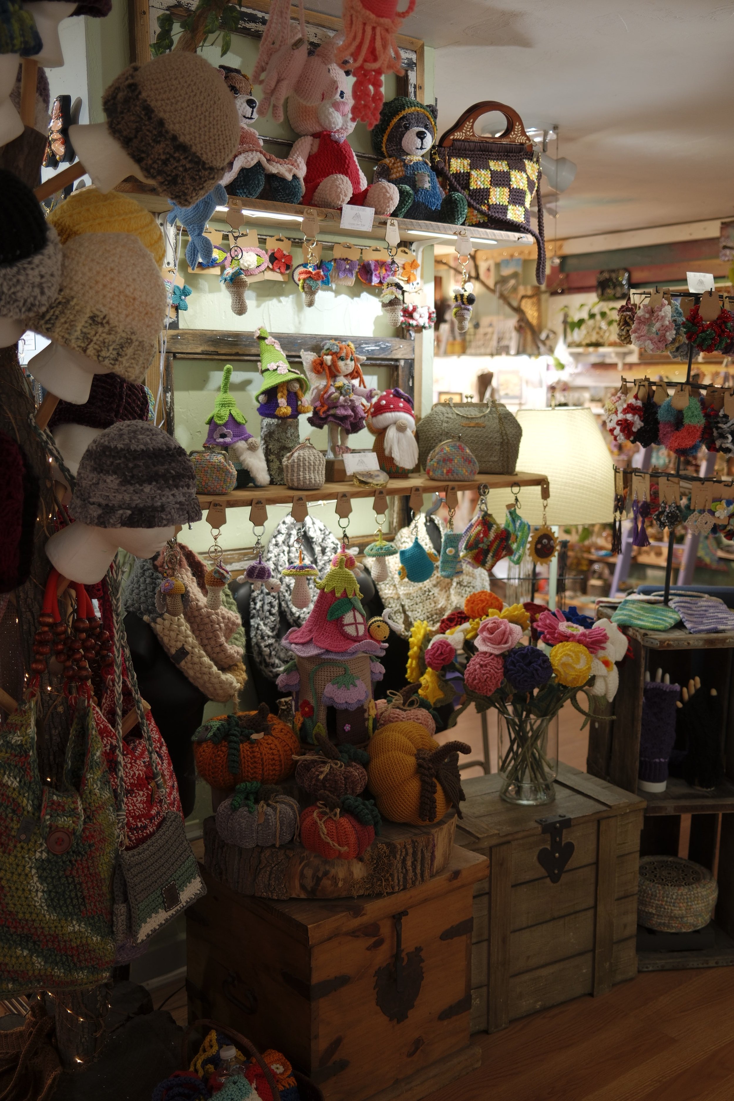 A display of handmade knitted and crocheted items including stuffed animals, hats, and accessories, arranged on wooden tables and shelves in a craft or gift shop.