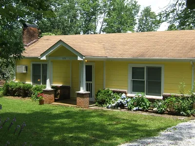 Yellow house with white trim, front porch with brick pillars, surrounded by green lawn and bushes, with trees in the background.