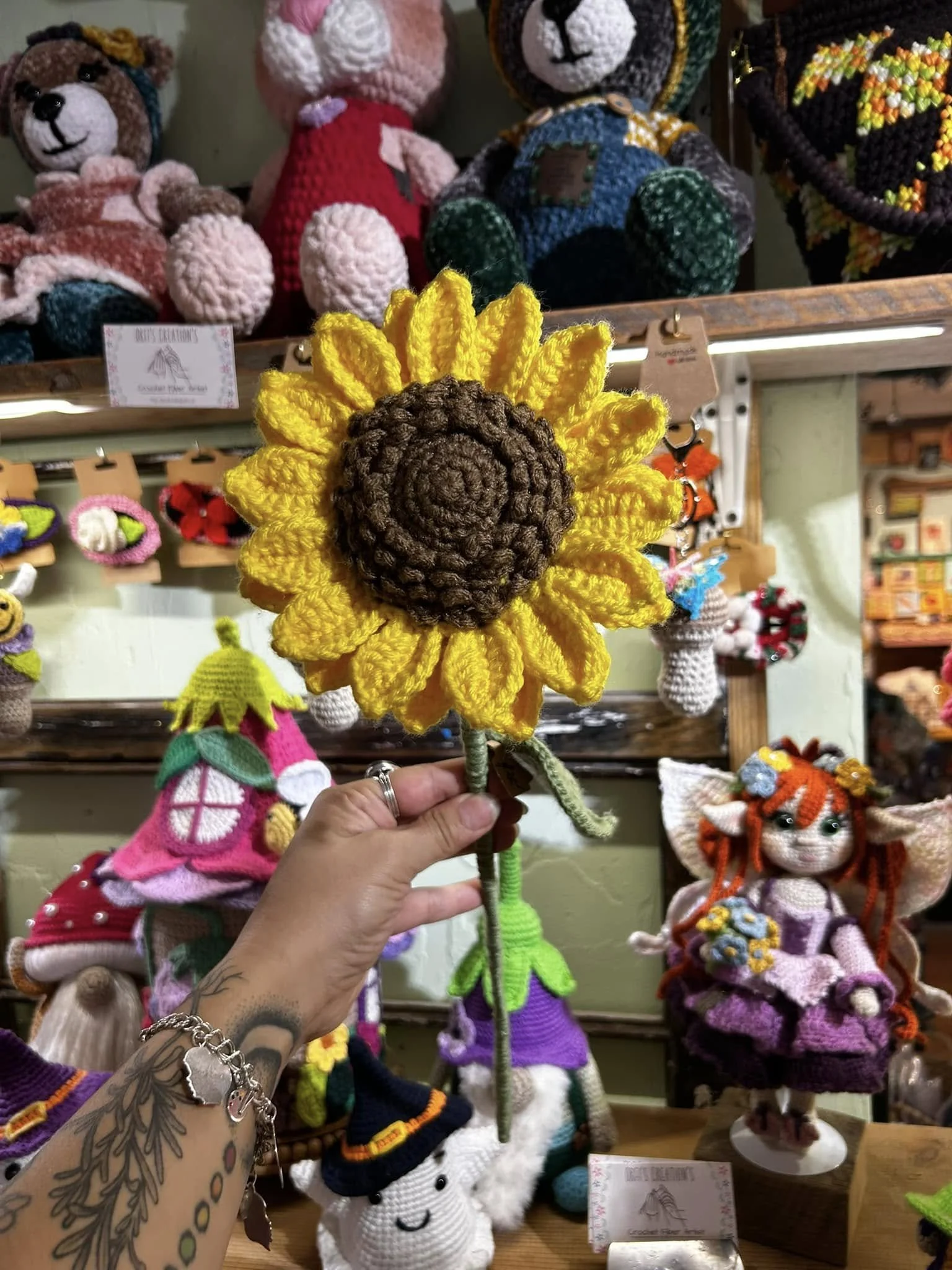 A person holding a yellow and brown crochet sunflower figure. The background features colorful various crochet dolls and figures, including animals and characters, displayed on shelves.