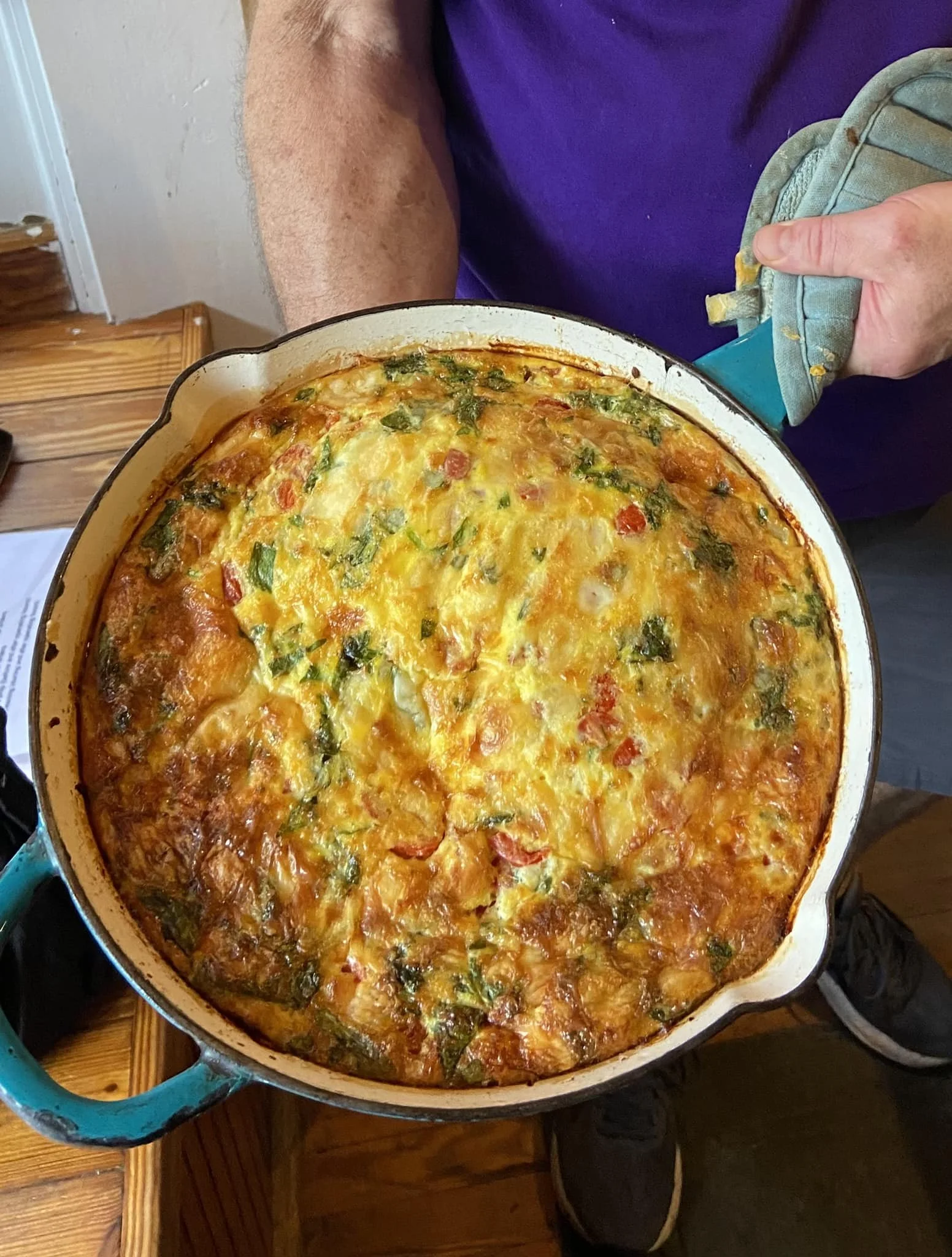 A person holding a skillet of homemade vegetable frittata, showing a colorful mixture of eggs, greens, and vegetables.