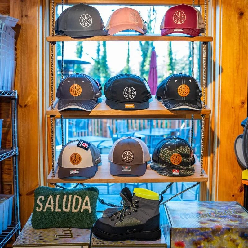 Display of various baseball caps on wooden shelves, in black, beige, red, navy, gray, and camouflage patterns, with a green knitted pillow reading "SALUDA" and a pair of black and yellow hiking boots on a table in front, in a store with outdoor scene