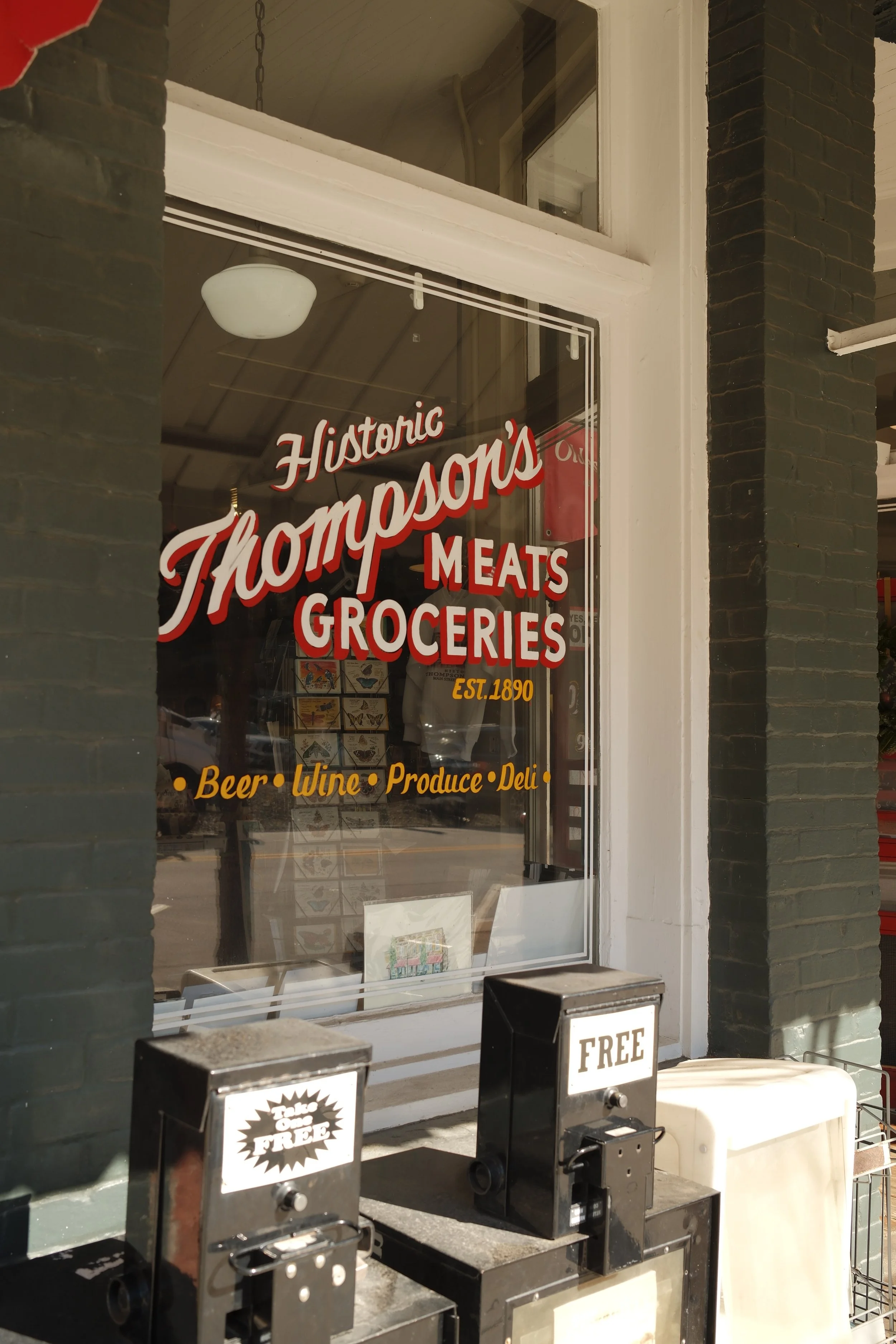 A storefront window for Thompson's, a grocery store established in 1890, displaying signs for groceries, meats, beer, wine, produce, and deli. There are also vending machines in front of the window offering free items.