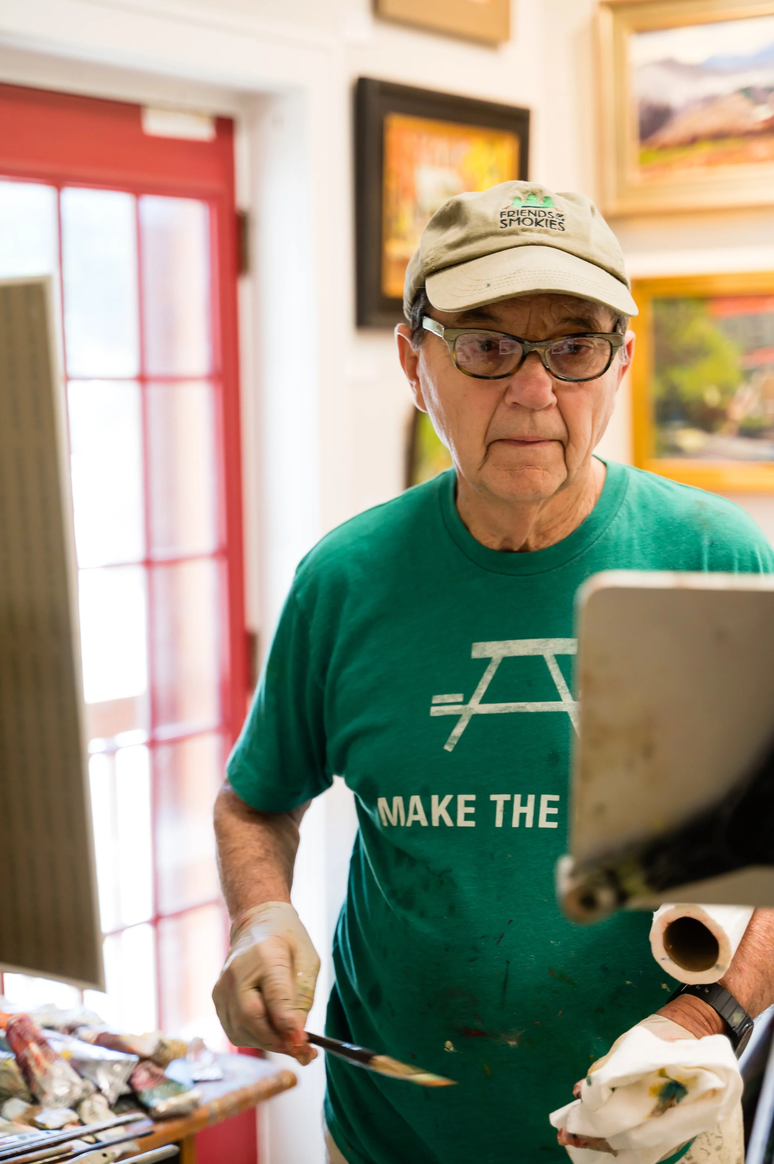 An elderly man wearing a beige cap with the words 'Friends of Smokies' and glasses, painting or working on an art project indoors. He is wearing a green T-shirt with white text and is holding a paintbrush and a paper towel.