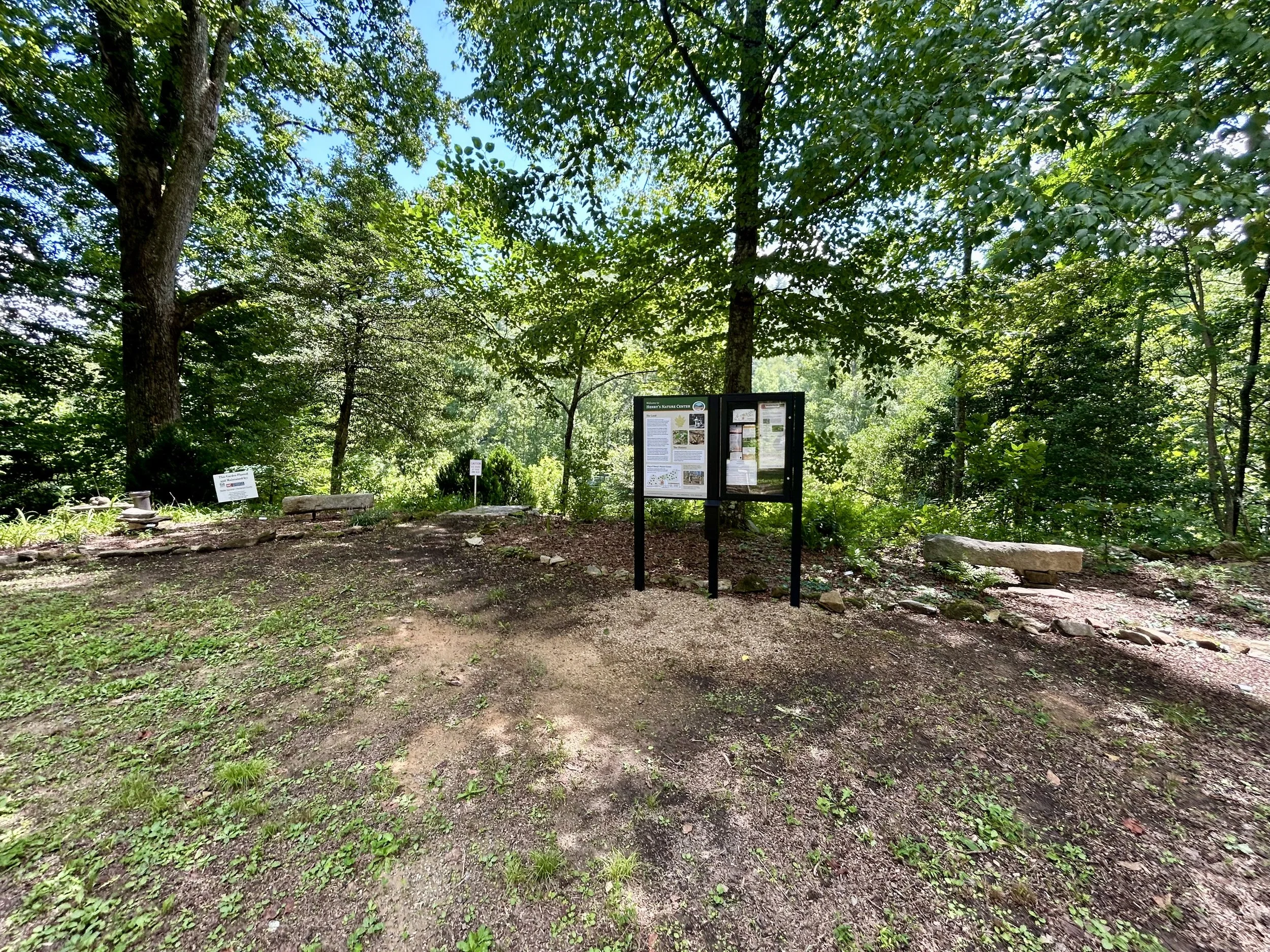 A wooded area with trees and green foliage, informational signs and benches, possibly part of a nature trail or park.