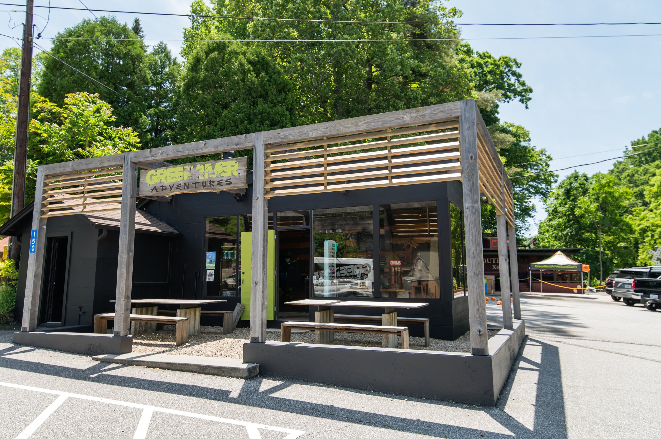 A small building with a sign that reads 'Green River Adventures' is visible in a parking lot with marked spaces and a few cars. The building has a black exterior, large glass windows, and is surrounded by green trees and a bright blue sky.