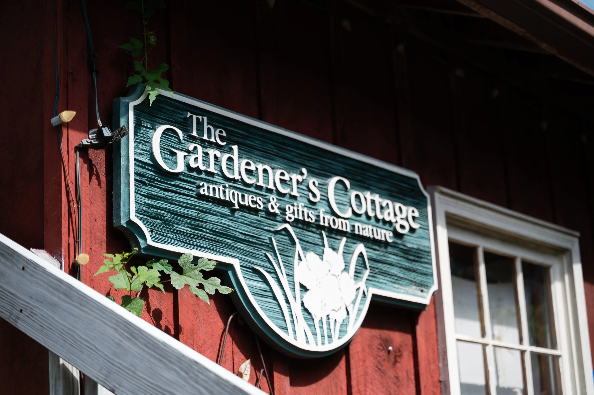 A sign for The Gardener's Cottage antique and gift shop mounted on a red wooden wall with some green leaves nearby.