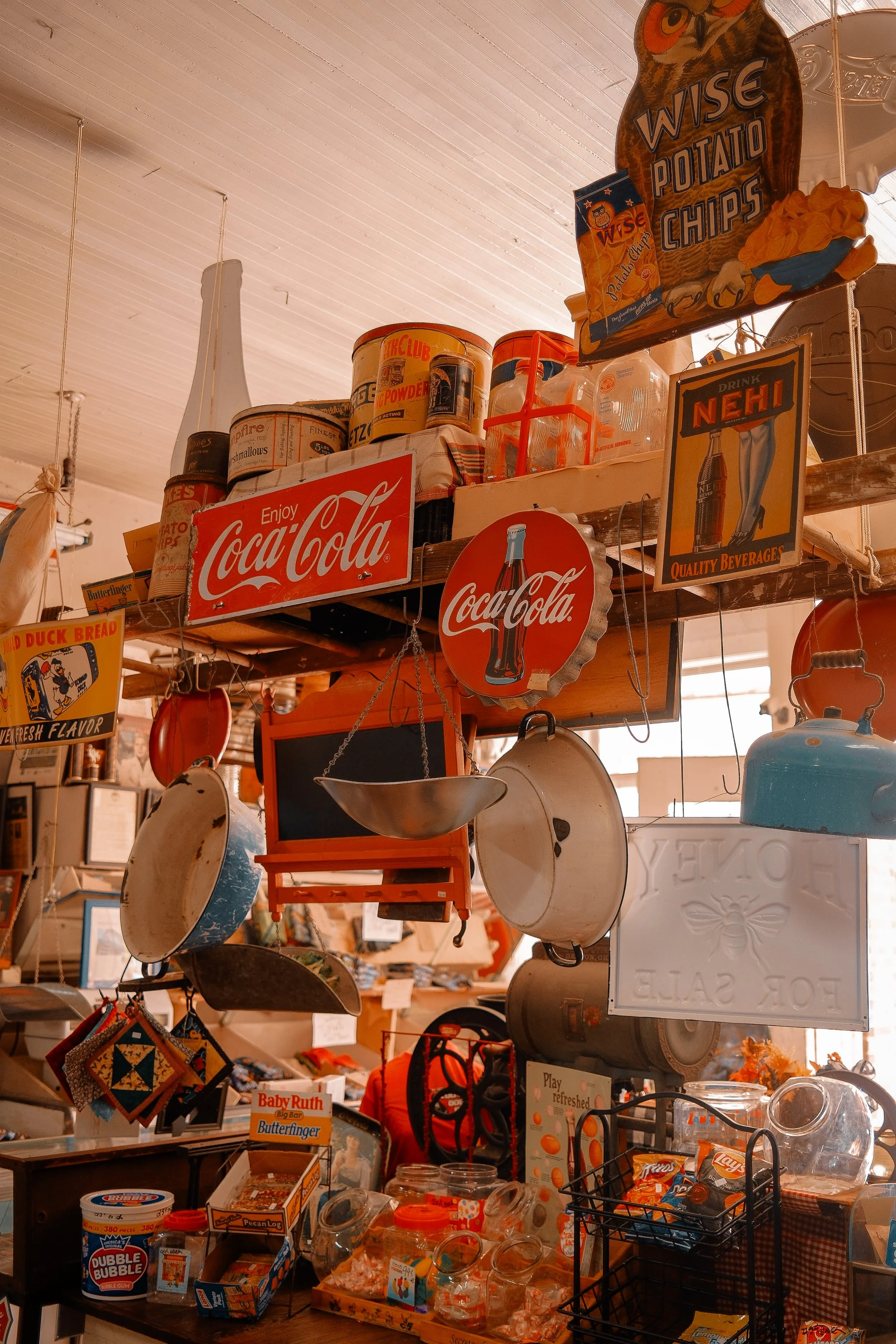 A collection of vintage and retro kitchen and grocery items on shelves, including pot pans, vintage Coca-Cola signs, snack bags, and jars, with a warm, nostalgic atmosphere.
