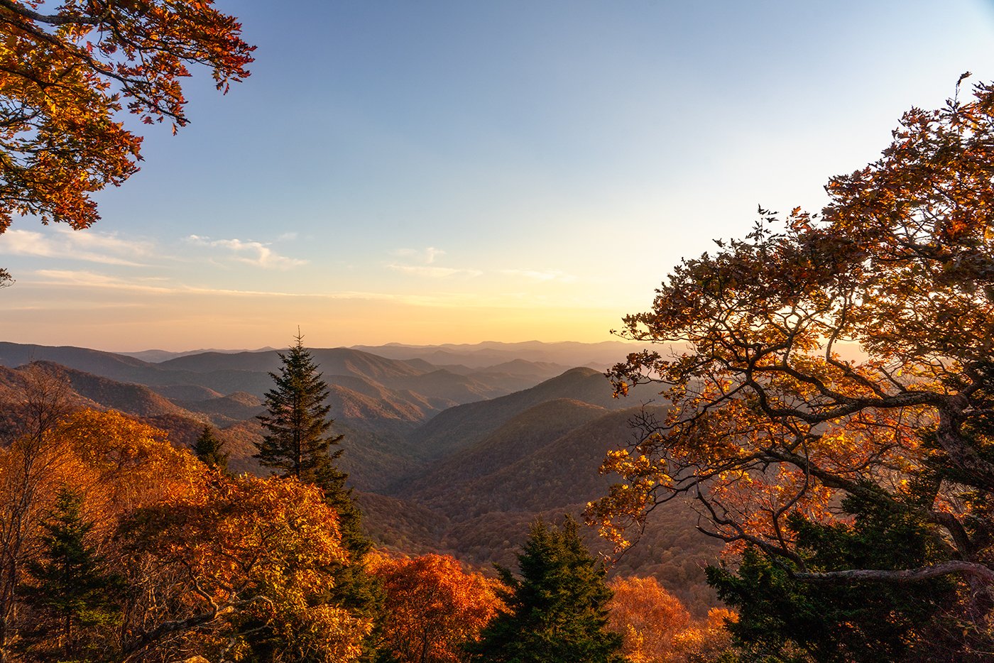 A scenic view of a mountain range during sunset with colorful autumn foliage in the foreground, including orange, yellow, and red leaves and a few green trees.
