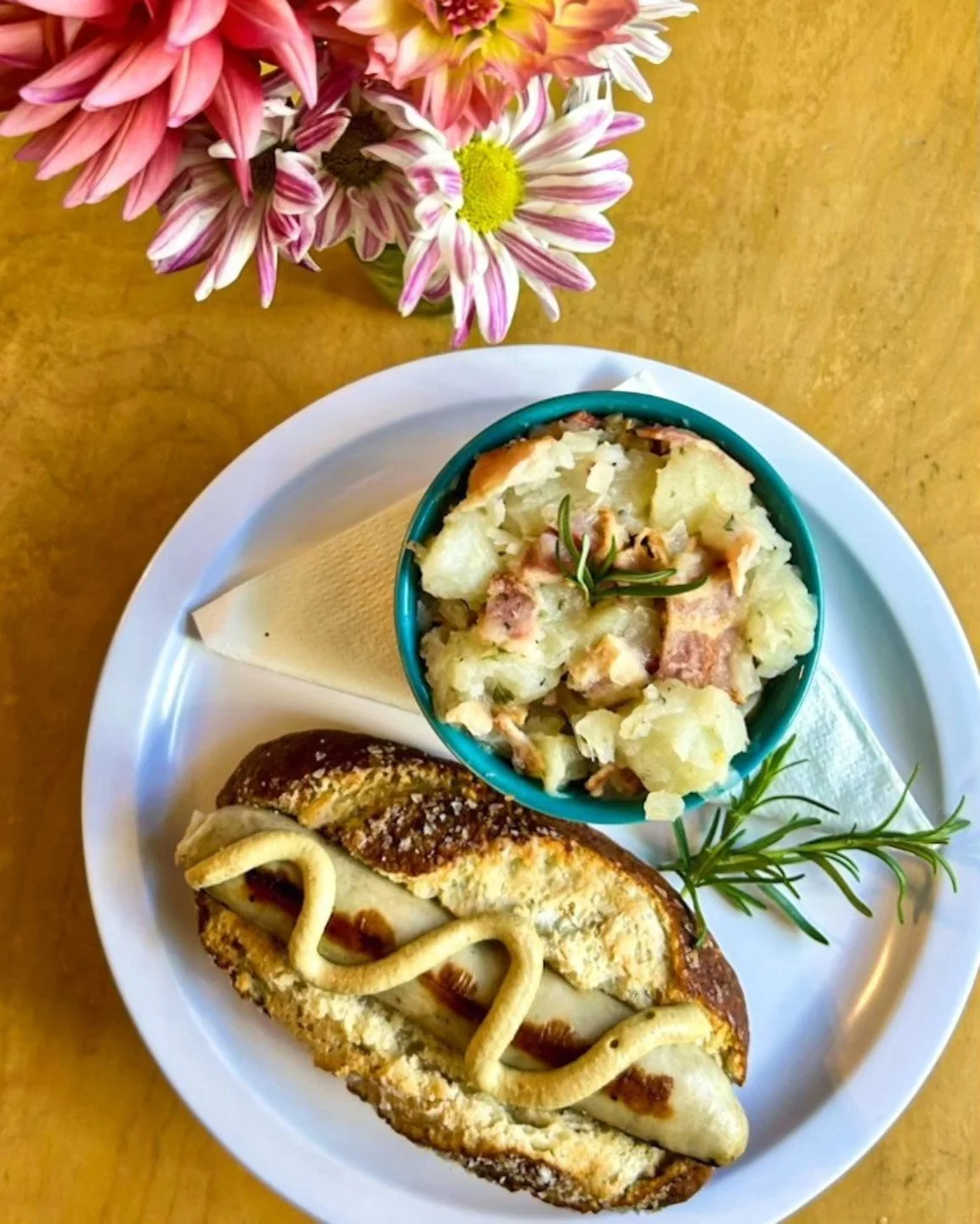 A plate with a hot dog topped with mustard, a small bowl of potato salad garnished with herbs, and a slice of cheese and cracker.