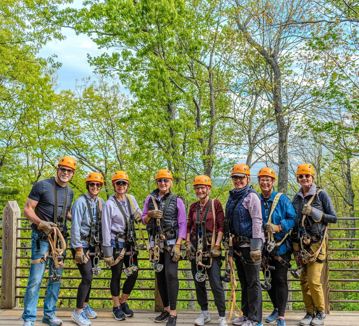 Group of nine people wearing safety helmets and harnesses on a wooden platform in a forest.