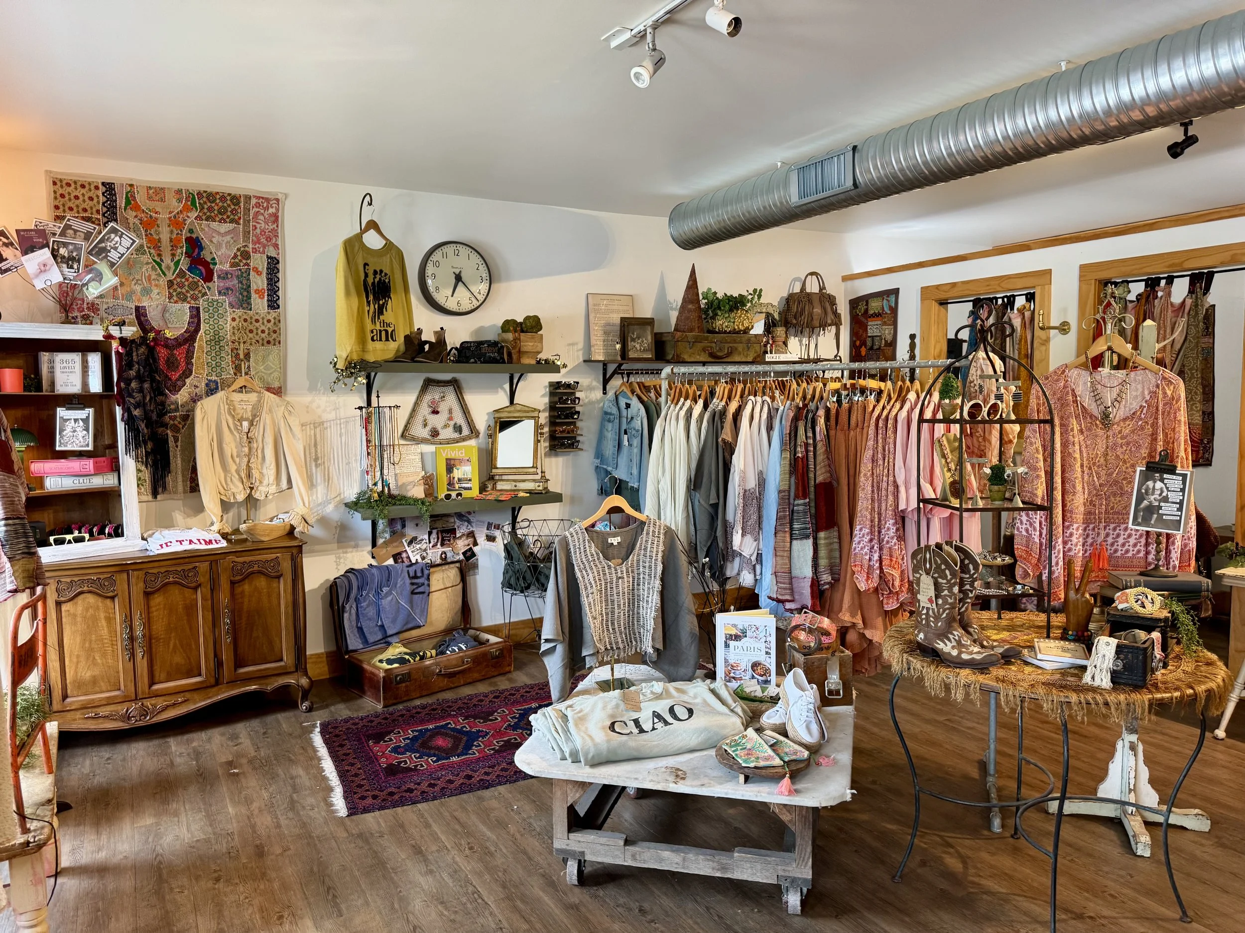 Interior of a vintage boutique clothing store with racks of clothes, shelves with accessories, and decorative items including a colorful patchwork wall hanging, a large wall clock, and framed pictures.