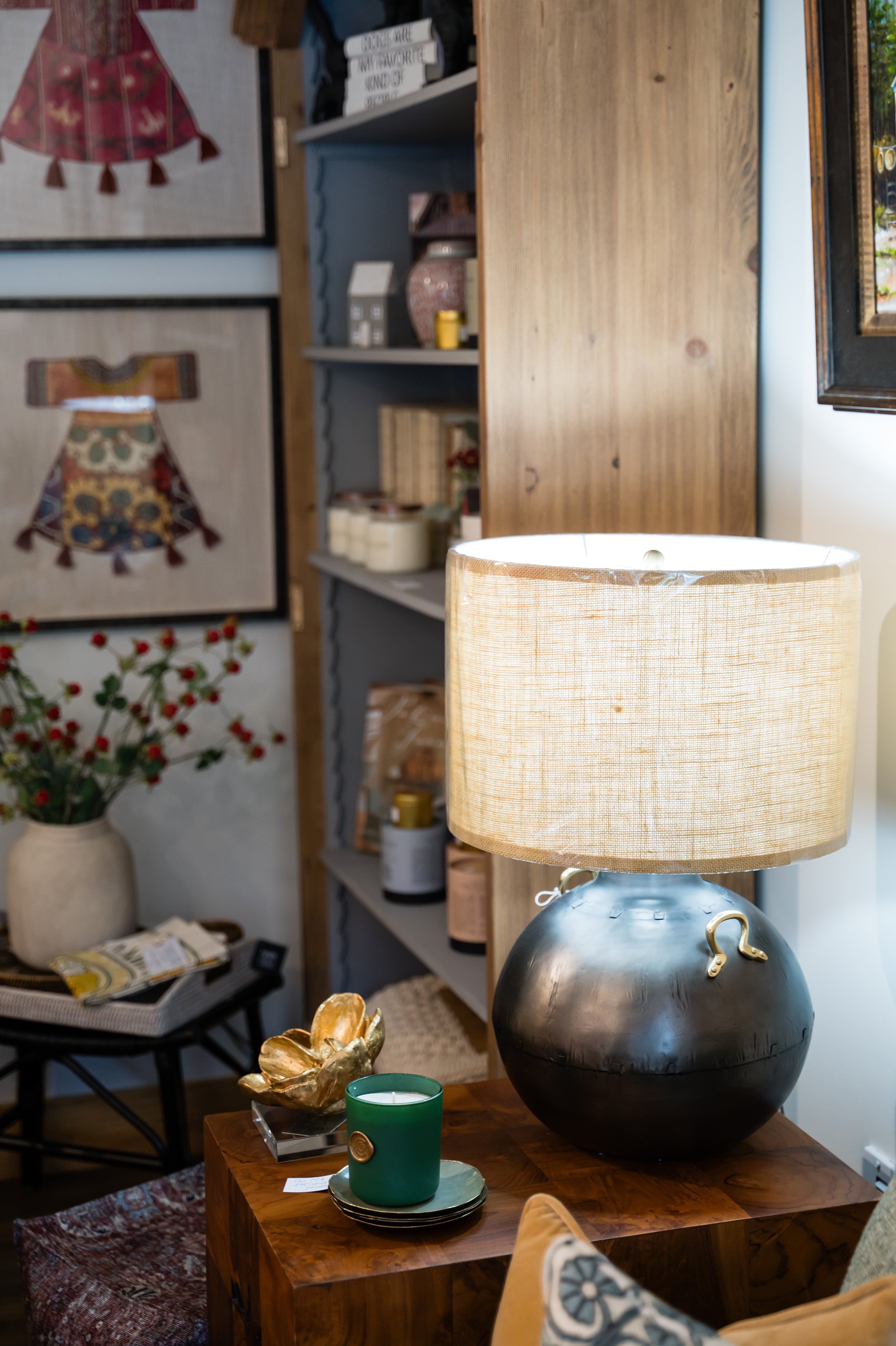 A cozy living room corner with a wooden side table holding a large black vase-shaped lamp with a beige drum shade, a green candle holder with a candle, and a decorative gold flower sculpture. Behind, there are framed woven art pieces, a bookshelf wit