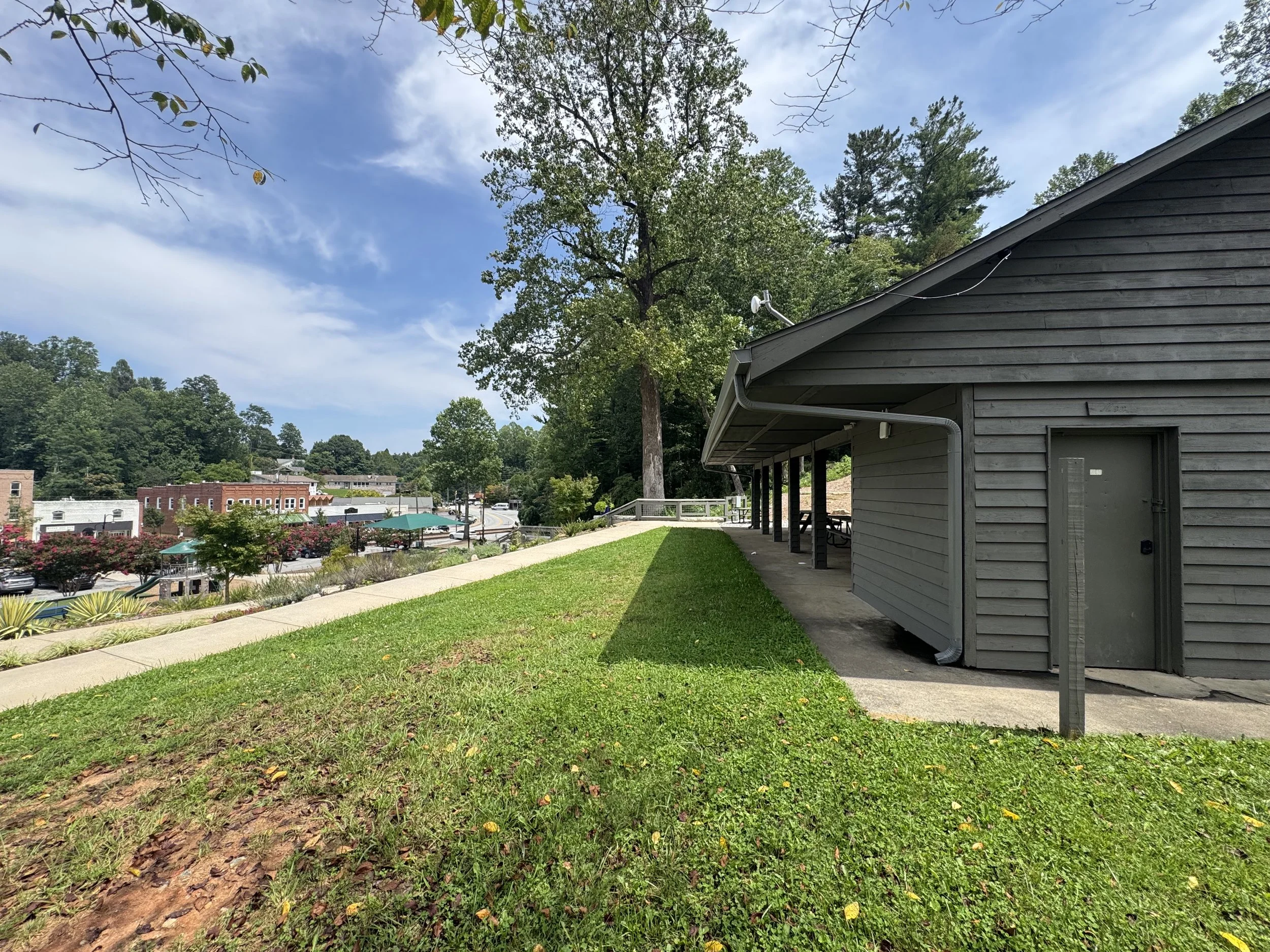 A small gray building with a sloped roof and a gutter, surrounded by green grass and trees, in a suburban area with a town visible in the background.