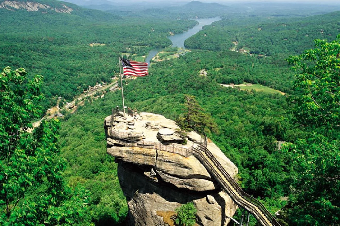 A rocky cliff with a staircase and an American flag on top, overlooking a green forest and a river in the distance.