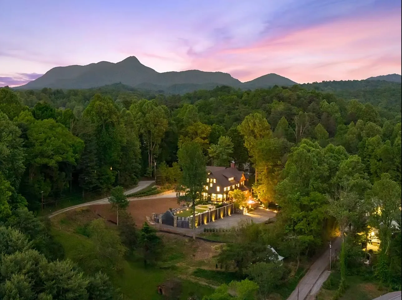 A house illuminated at dusk surrounded by a lush forest with mountains in the background and a colorful sky