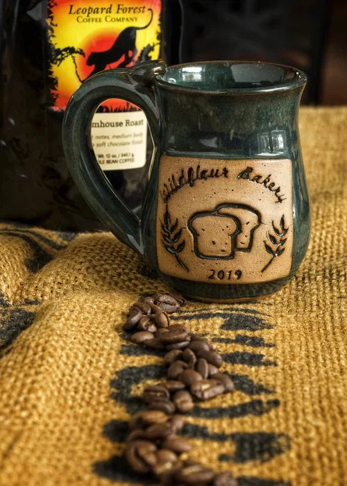 A ceramic mug with a greenish glaze and tan front panel that has a bread and brain illustration with the text 'Mindful Coffee' and the year 2019. Next to the mug are scattered coffee beans on a textured burlap surface. In the background, there is a b