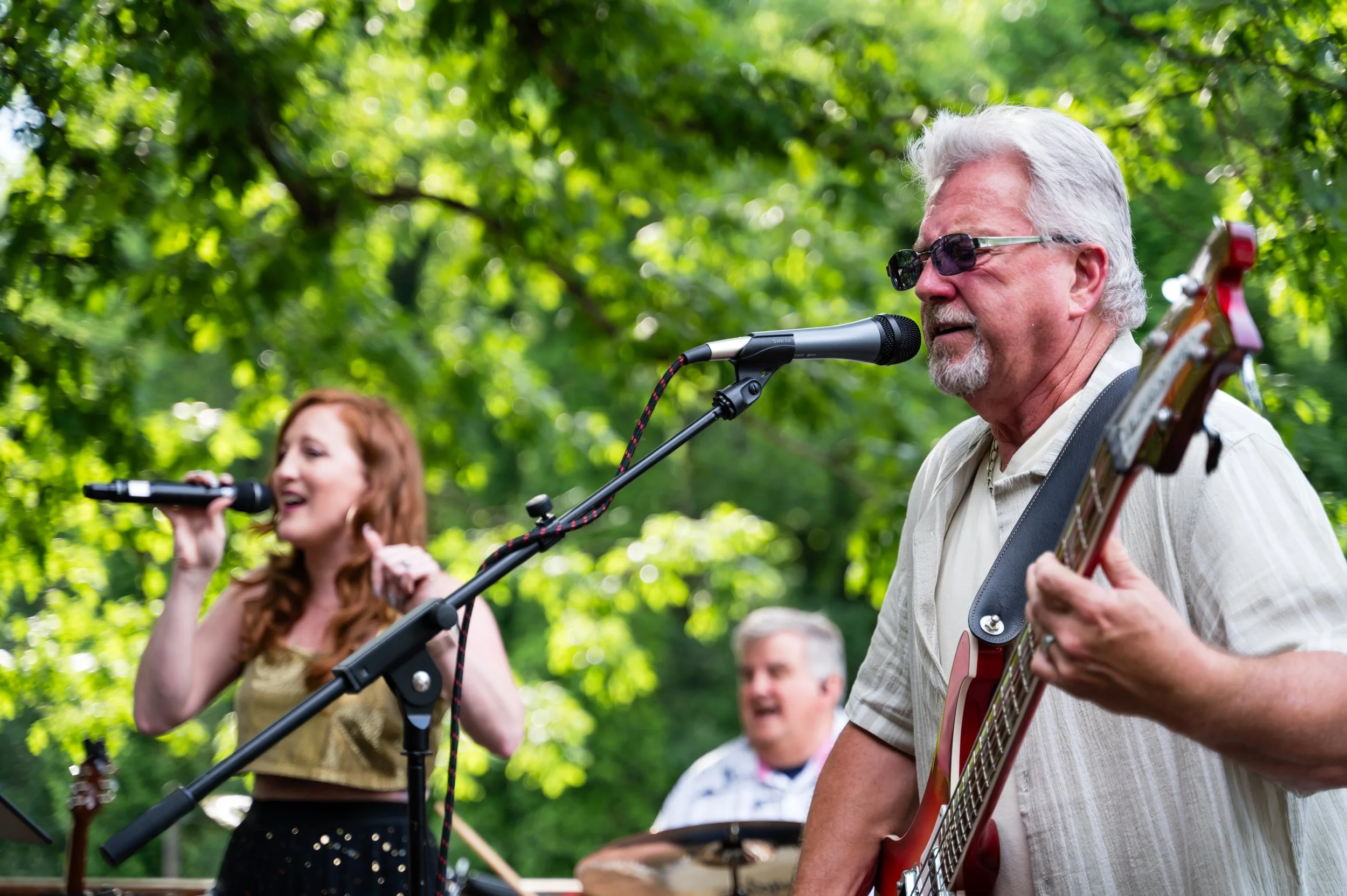 A group of people outdoors, with a man wearing sunglasses singing into a microphone and playing an acoustic guitar, while a woman next to him is also singing into a microphone.