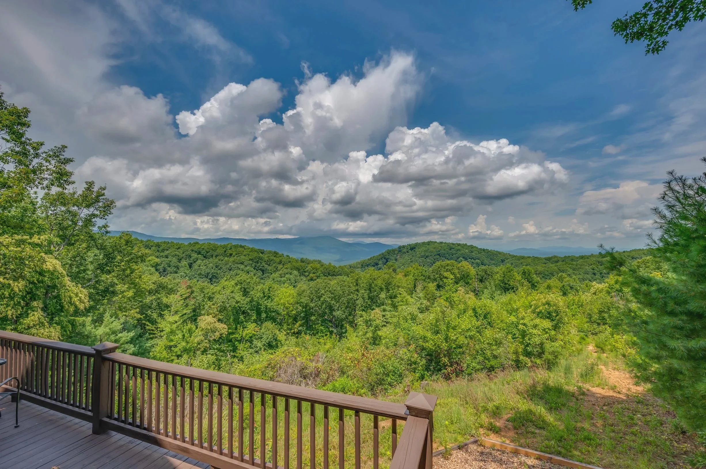 View of a lush green forested landscape with rolling hills under a partly cloudy sky, seen from a wooden deck with a railing.