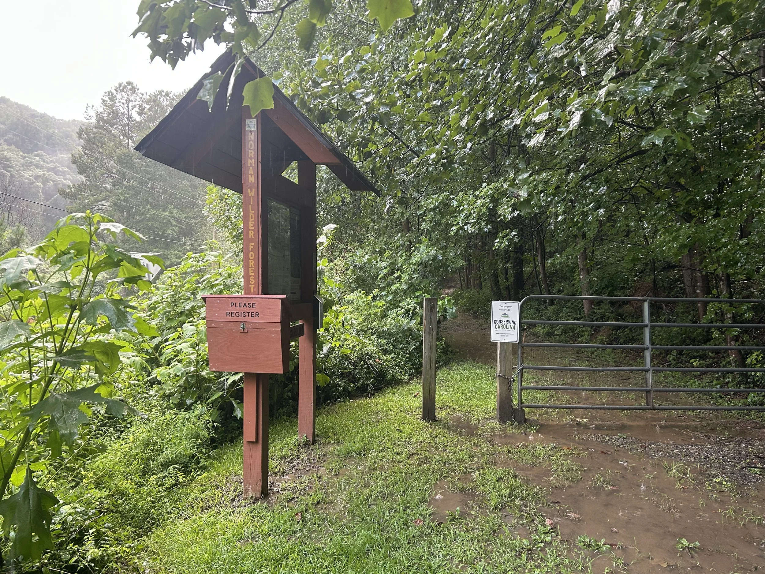 A small wooden registration kiosk with a red vertical sign and a pink box labeled 'Please Register,' situated near a metal gate in a wooded area with lush green trees and foliage.