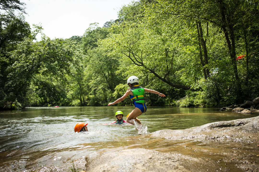 Three children playing in a river surrounded by lush green trees, with one child jumping over a rock wearing a helmet and life jacket, and the other two children swimming nearby.