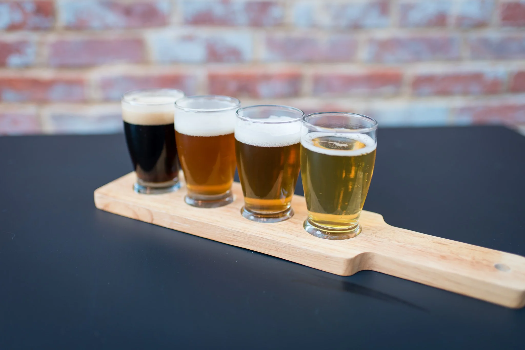 A flight flight presentation with five glasses of beer in a row on a wooden tray, set against a background of exposed brick wall.