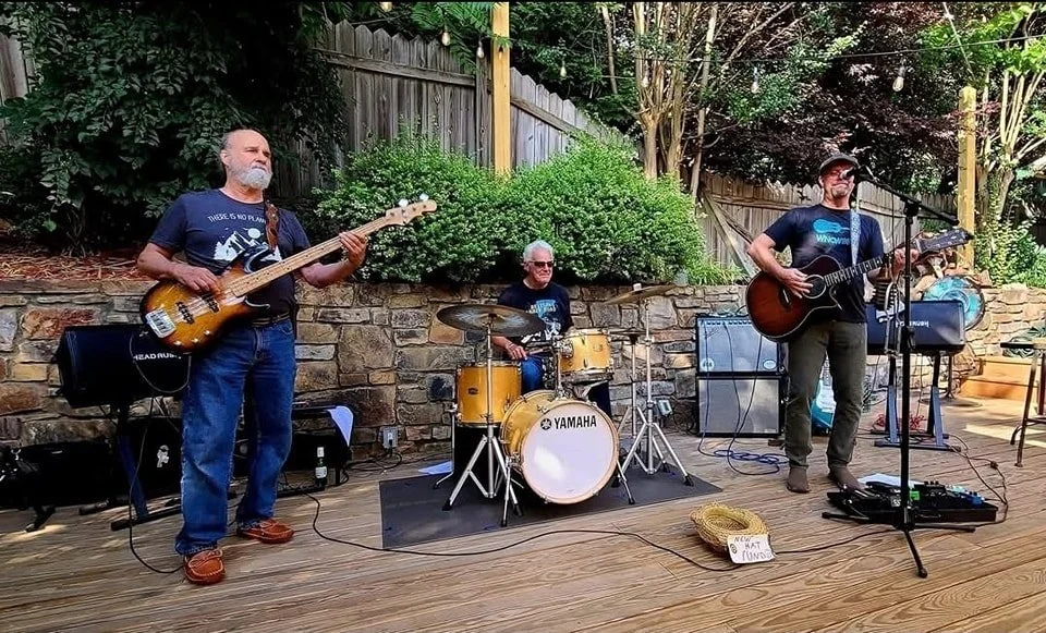 Three musicians performing outdoors on a wooden deck with a brick wall, green bushes, and trees in the background. One man is playing a bass guitar, another man is playing drums, and a third man is singing and playing an acoustic guitar.