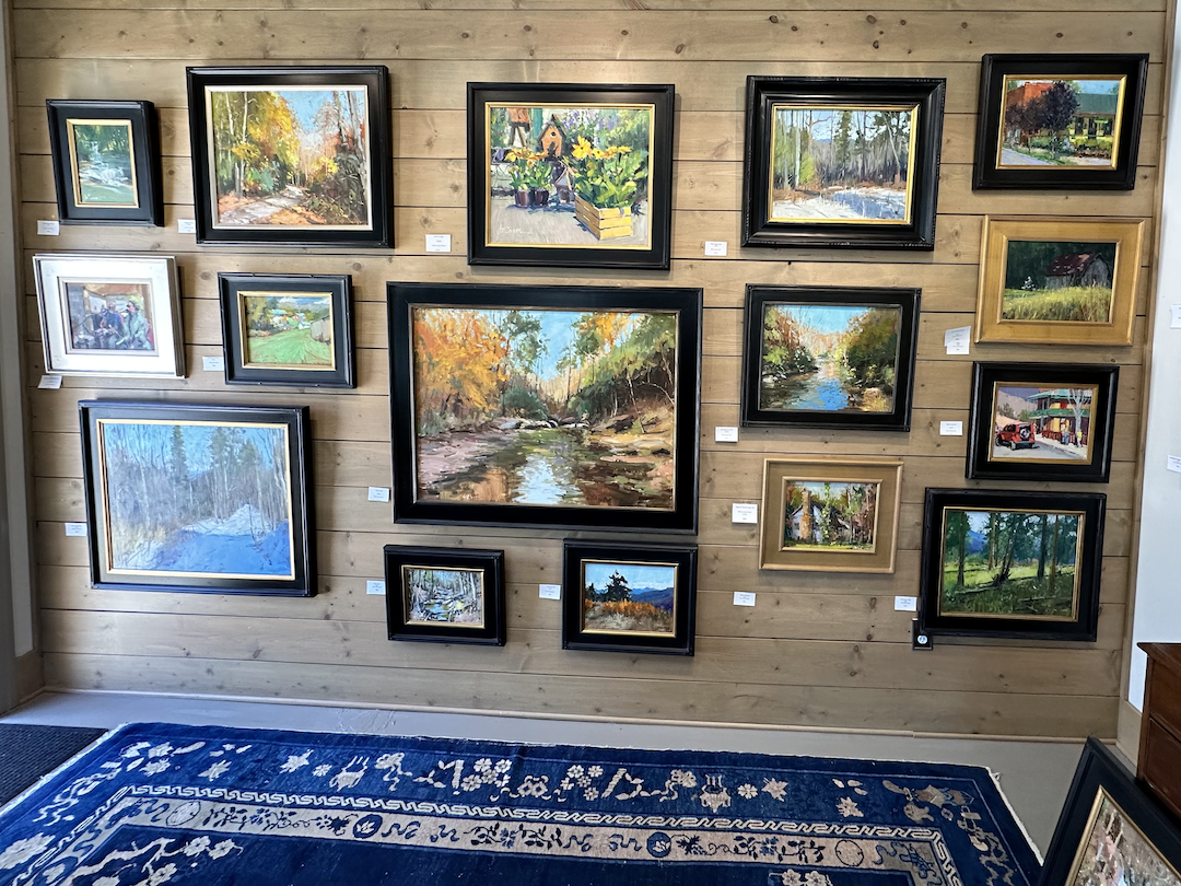 Wall display of various landscape paintings in different sizes and frames, mounted on a wooden wall with a blue patterned rug in front.