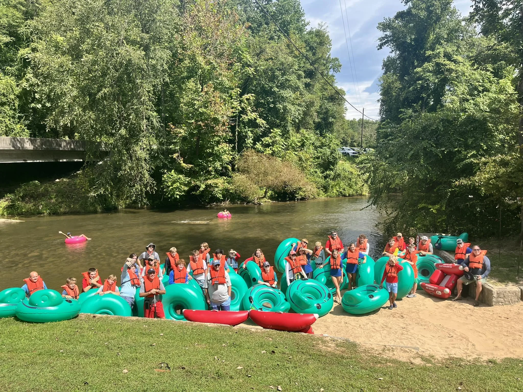 Group of children and adults gathering by a river, preparing for a kayaking trip, with many teal kayels, life jackets, and inflatable tubes. Dense green trees line the riverbank under a partly cloudy sky.