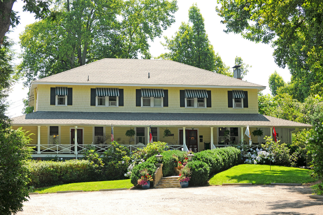 A large yellow two-story house with a wraparound porch decorated with hanging plants, flags, and outdoor furniture, surrounded by a well-maintained garden with bushes, flowers, and a gravel driveway, set in a lush green environment.