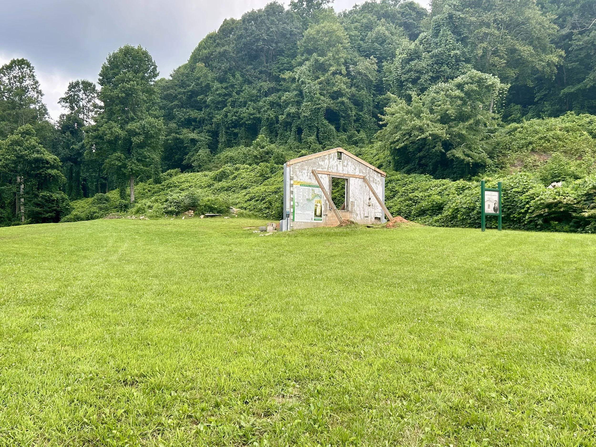 A small, weathered building with a building information sign in front, set in a lush green landscape with dense trees on a cloudy day.