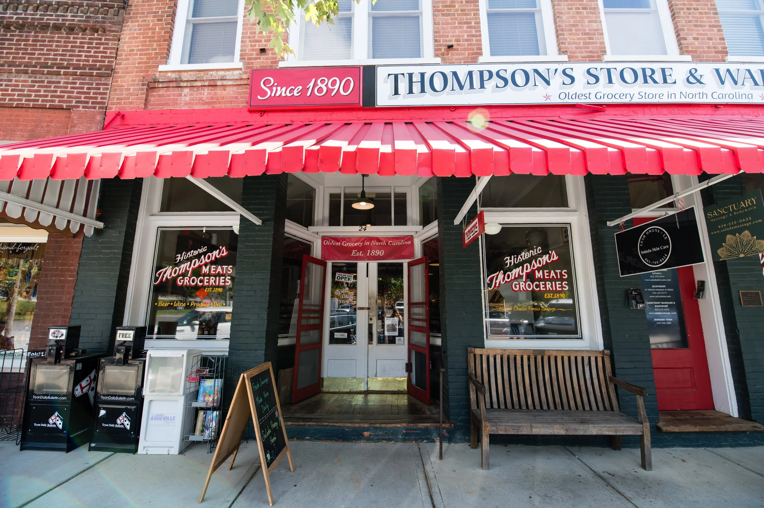 The storefront of Thompson's Store & W in North Carolina, established in 1890, featuring a red and white striped awning, brick facade, and windows. The entrance is open, with signs advertising groceries, meats, and other products. There is a sidewalk