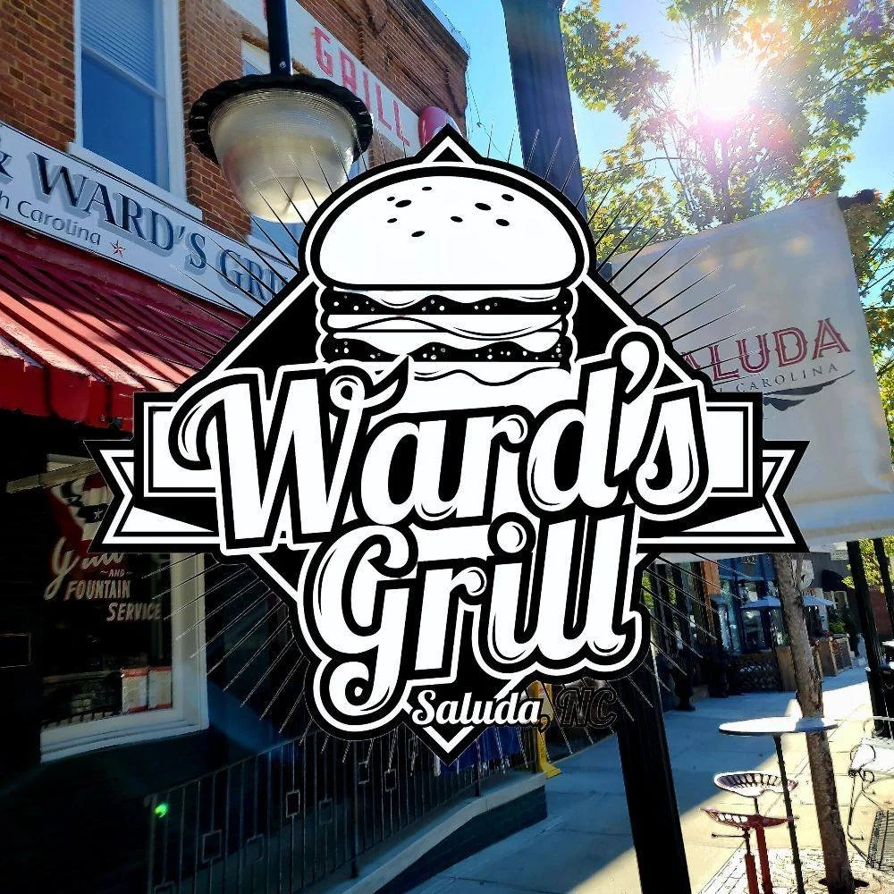 Sign for Wards Grill in Saluda, NC with a graphic of a hamburger on it, hung on an outdoor storefront amidst trees and sunlight.