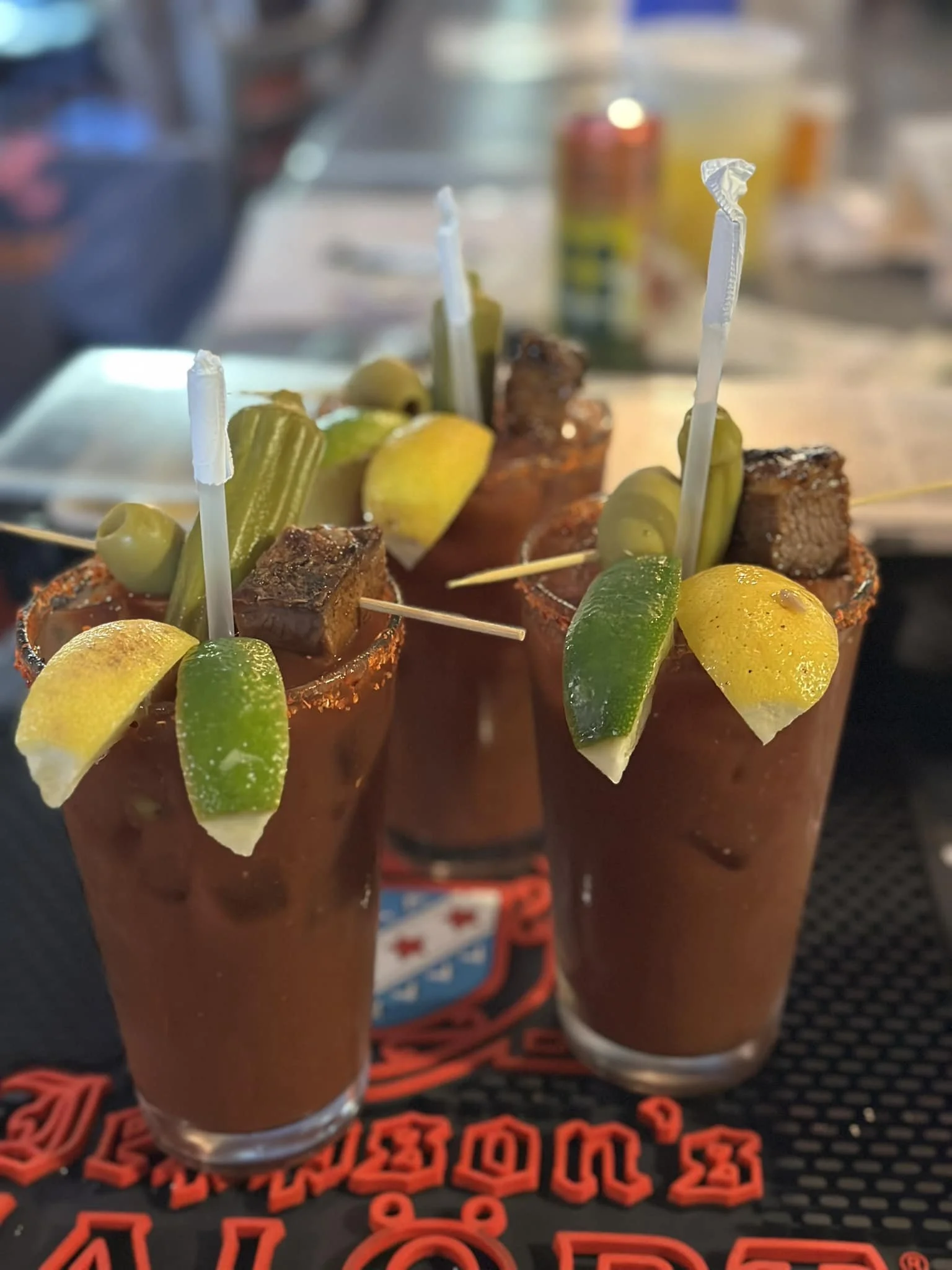 Three Bloody Mary drinks garnished with lemon wedges, lime wedges, pickles, olives, and small pieces of cooked meat with white stir sticks, placed on a black and red textured surface.