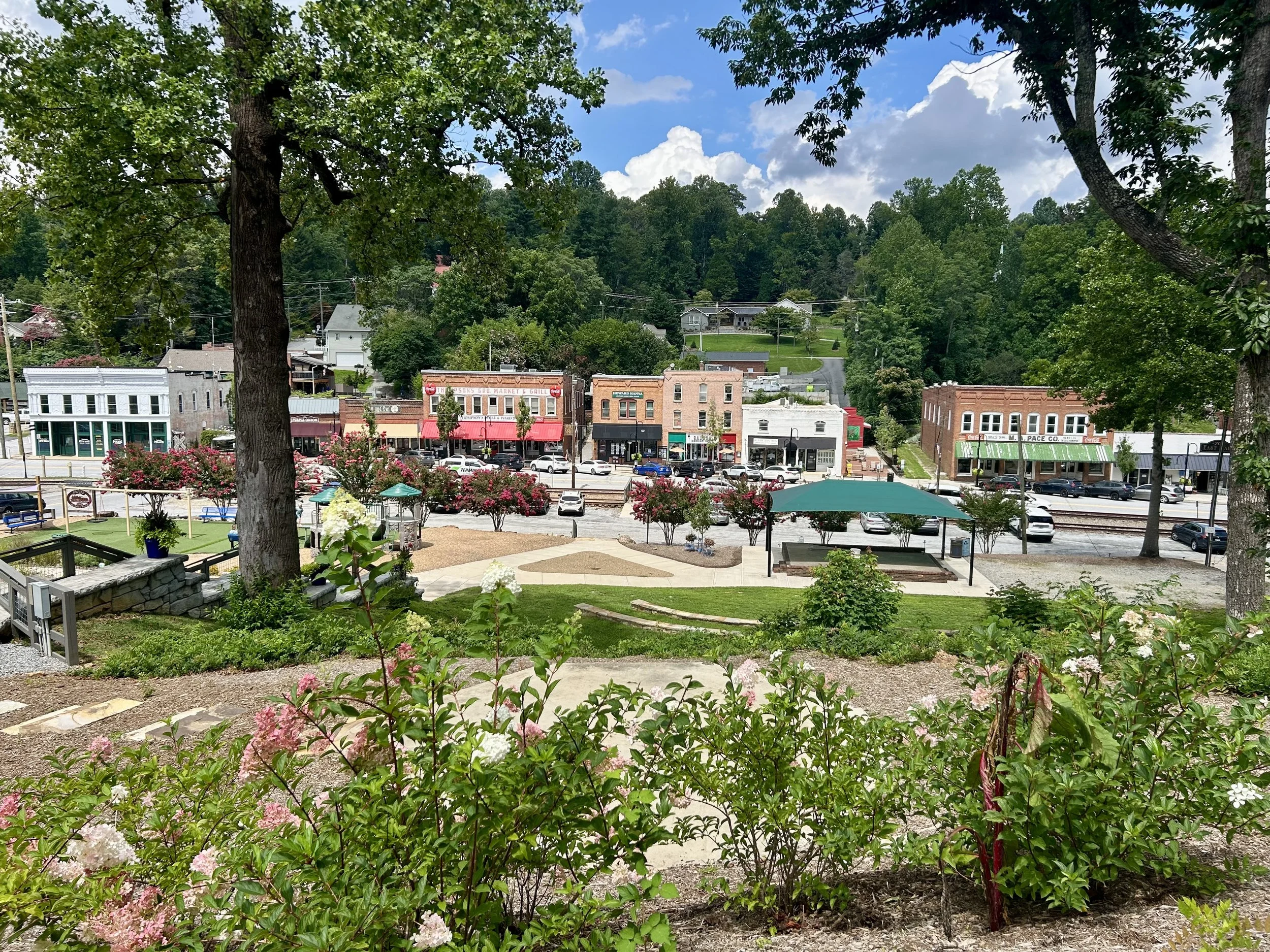 A picturesque view of a small town or village with storefronts on the main street, surrounded by lush greenery, trees, and hills under a partly cloudy sky.