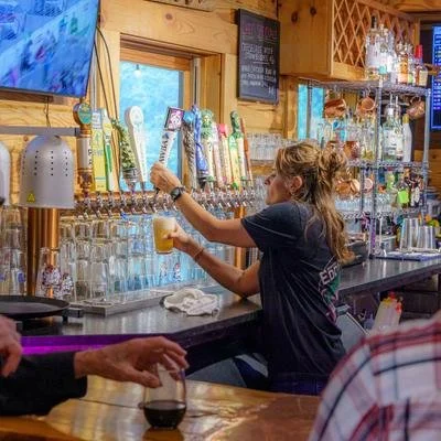 A woman with curly hair in a black shirt pours a draft beer at a bar with liquor bottles and beer taps in the background.