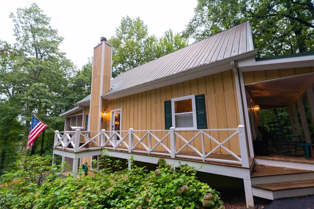 Yellow house with white porch railing, green shutters, and an American flag, surrounded by trees.