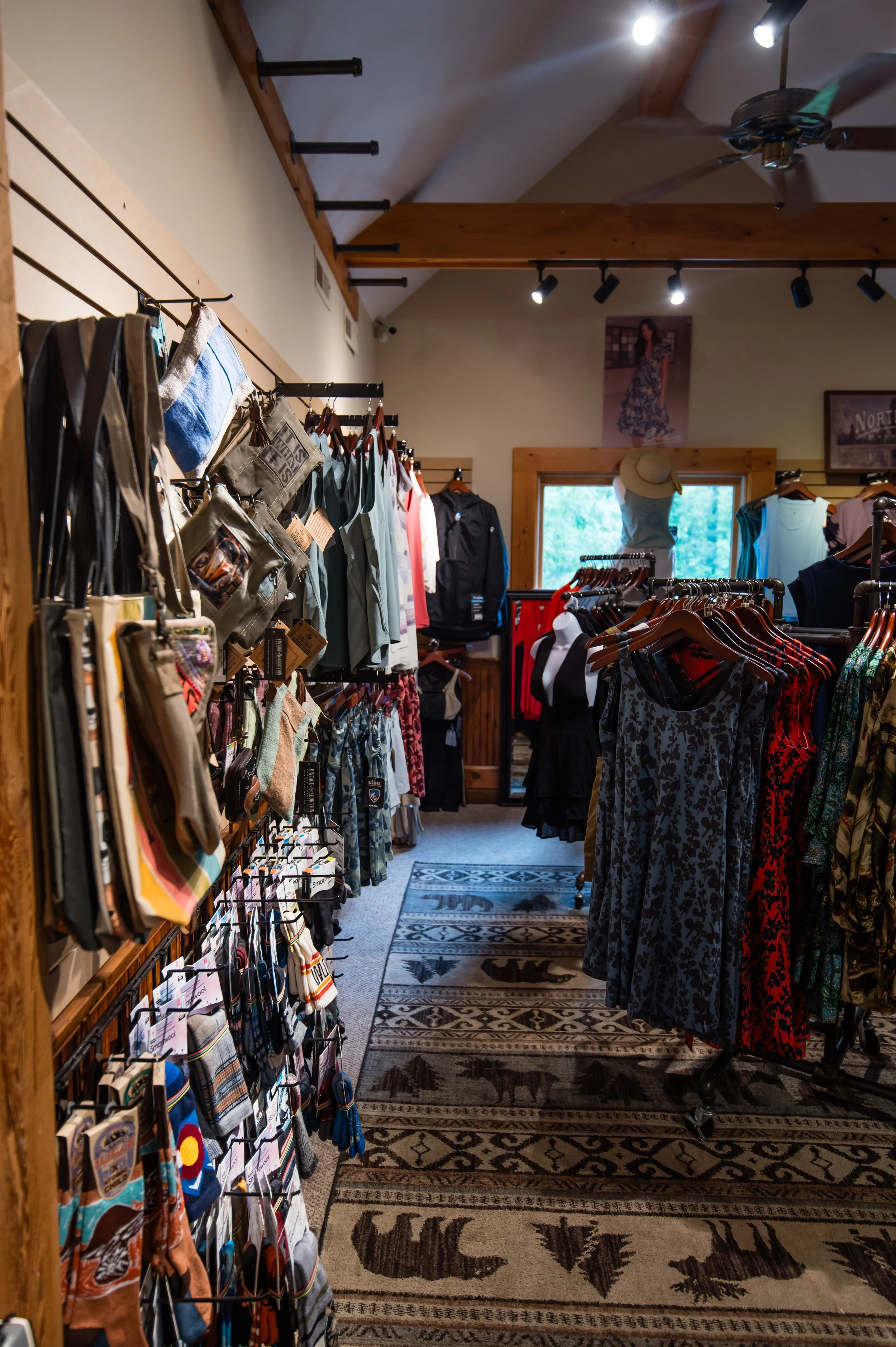 Inside a clothing store, displaying various apparel and accessories on racks and shelves, with wooden accents and a patterned rug on the floor.