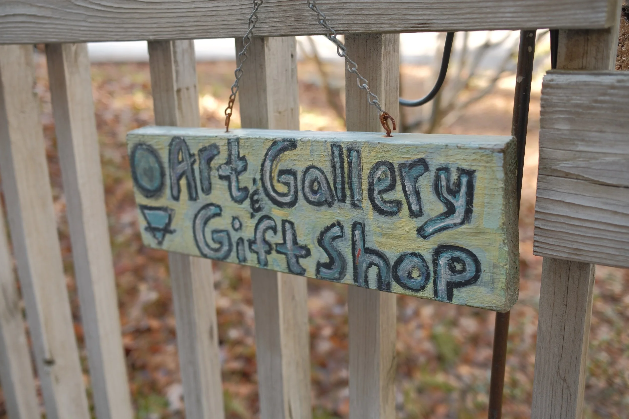 Close-up of a hanging wooden sign with hand-painted black letters, attached with a rusted chain, mounted on a wooden fence outdoors.