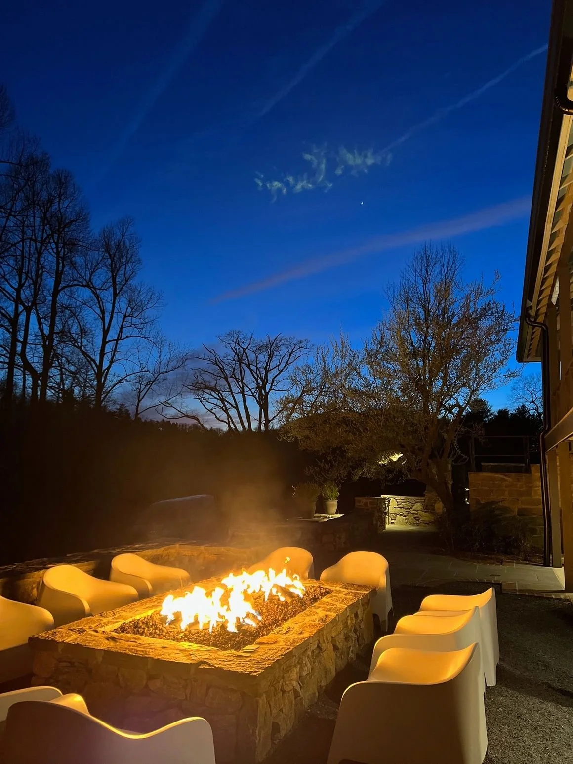 An outdoor seating area with a stone fire pit surrounded by white chairs, lit by a blazing fire against a night sky with trees and a house on the right.