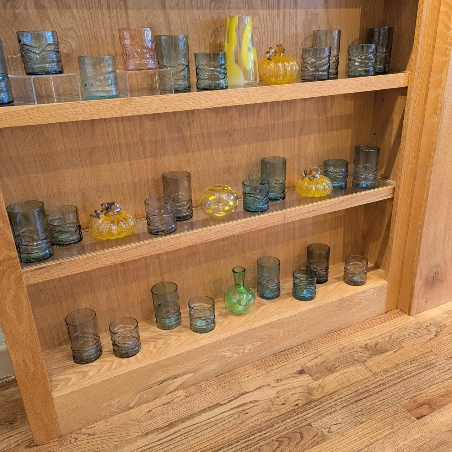 A wooden shelf filled with hand-blown glass cups, and decorative glass pumpkins in yellow and green.